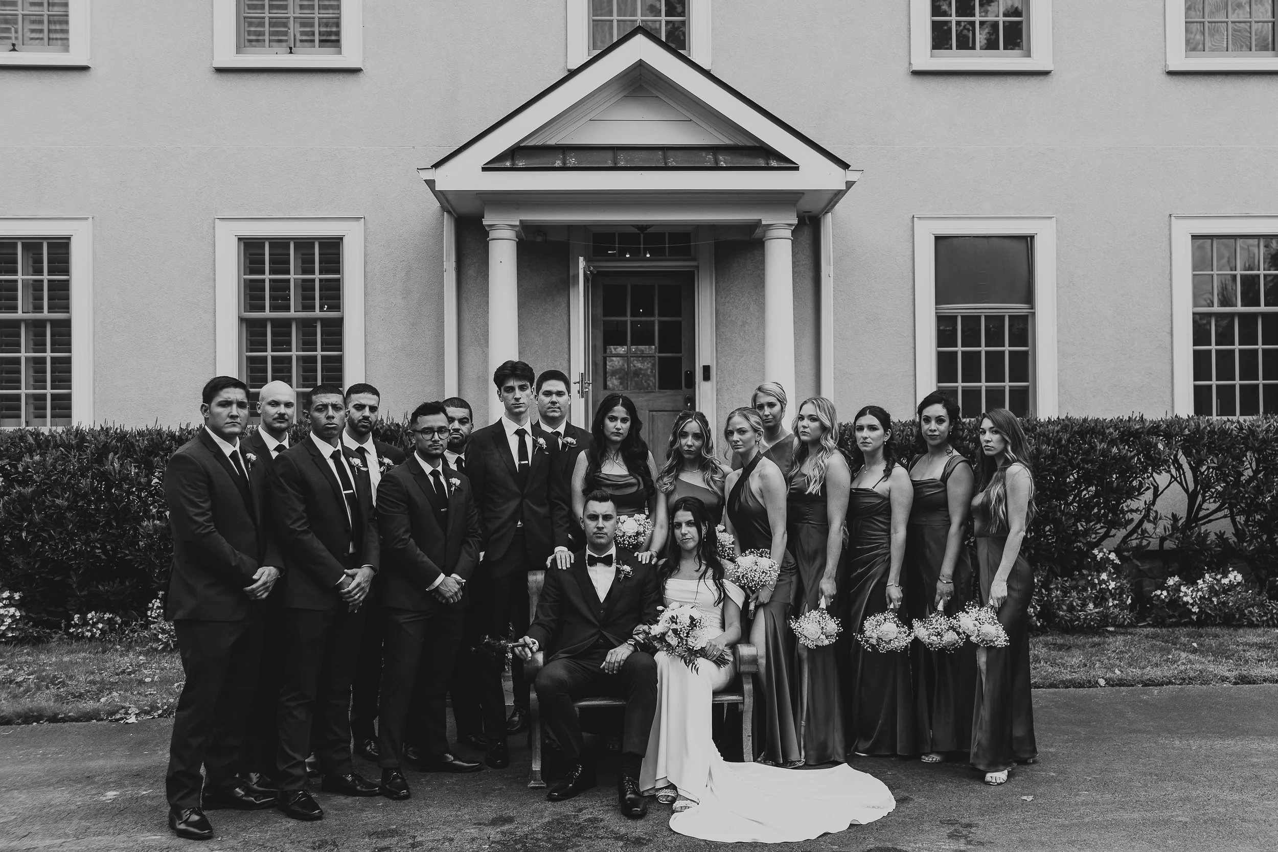 Black and white photo of a wedding party standing in front of a house. The group includes men in suits, women in dresses, and a bride in a white gown holding a bouquet, with a groom kneeling in front. The house has multiple windows and a porch with c