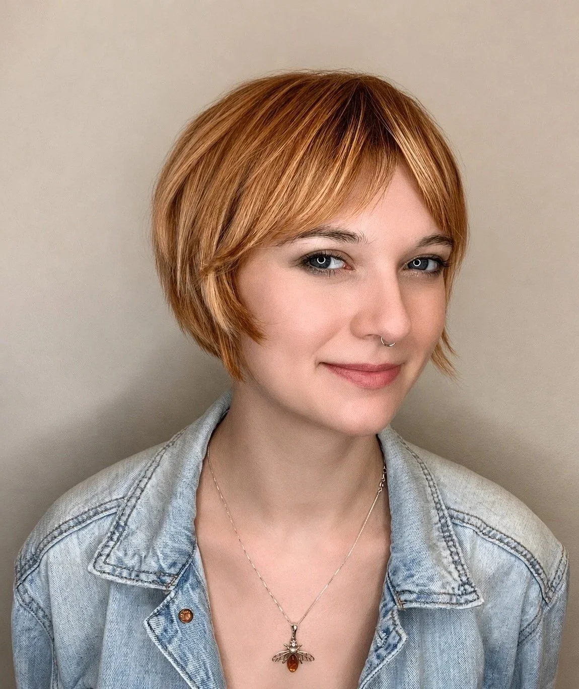 Young woman with short, layered, red hair, light makeup, and a septum piercing, smiling slightly, wearing a light denim jacket and a necklace with a bee pendant, standing against a plain neutral background.