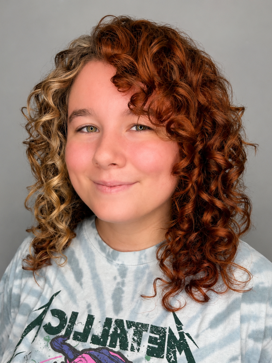 A young girl with light skin and curly hair, half blonde and half red, smiling softly, wearing a tie-dye t-shirt with a graphic design.