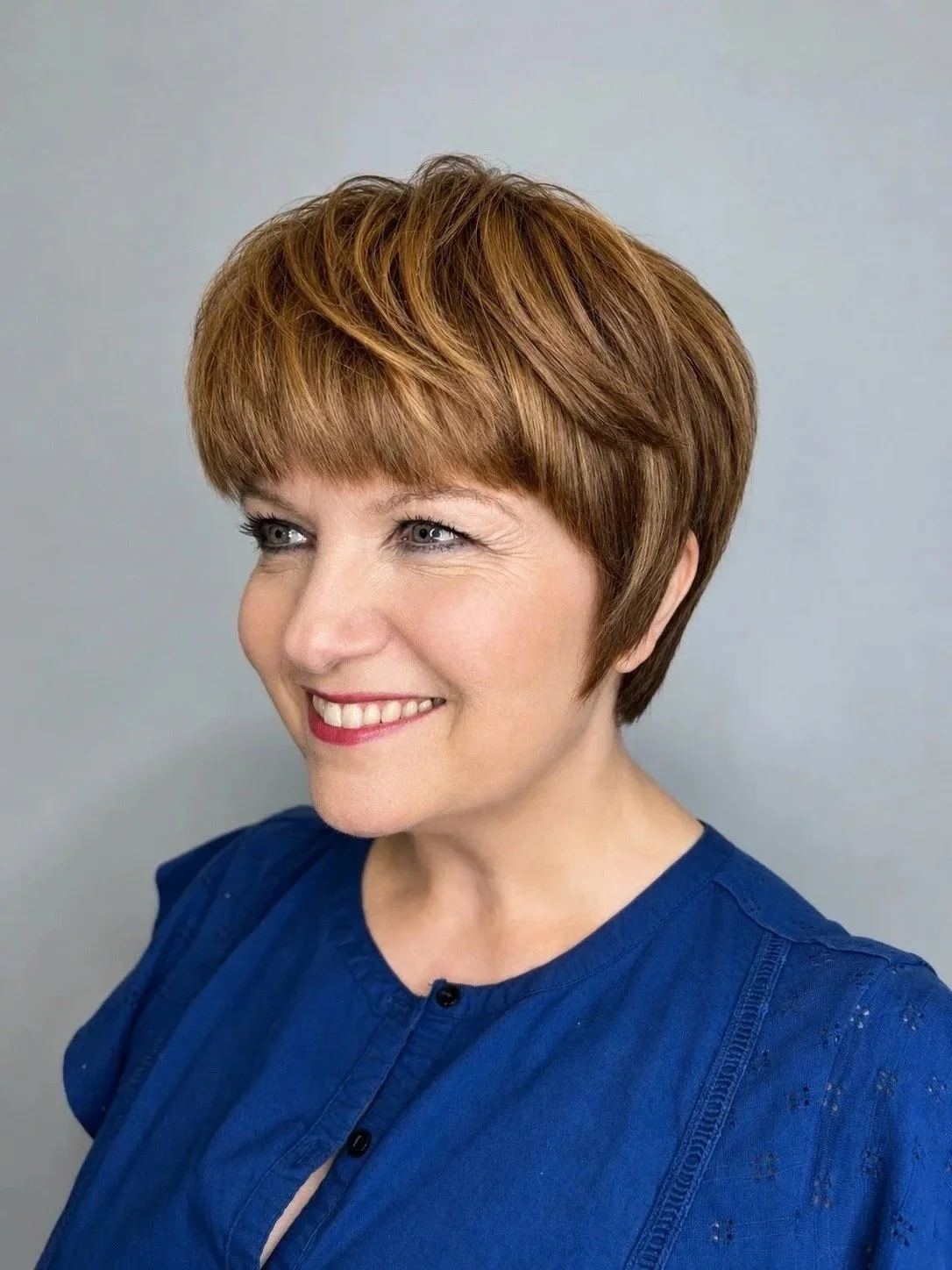 A woman with short, reddish-brown hair smiling, wearing a blue blouse, against a plain grey background.