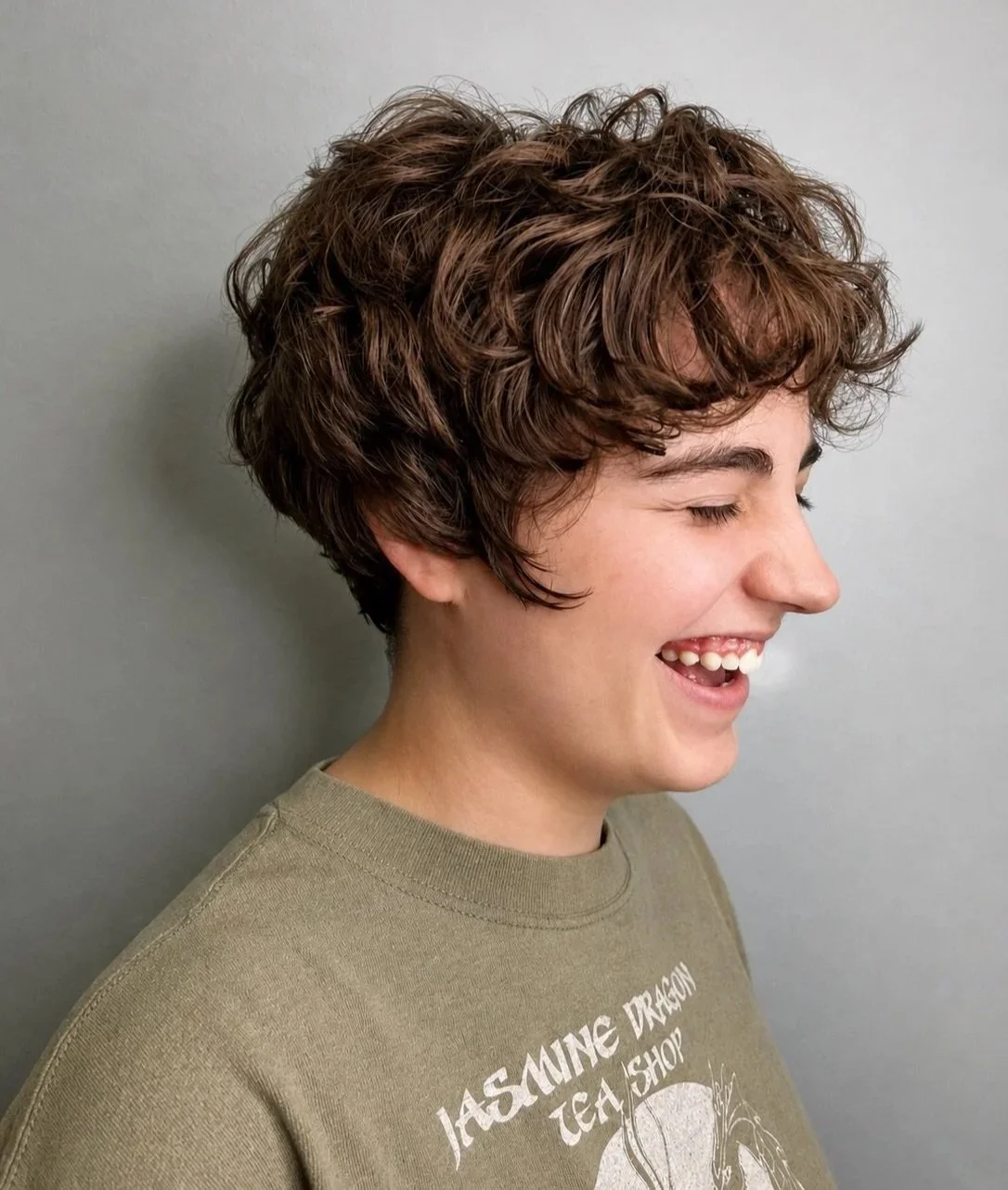 Side profile of a young person with short, curly brown hair, smiling with closed eyes, wearing a beige T-shirt with a JAS MINE DRAGON text and graphic.
