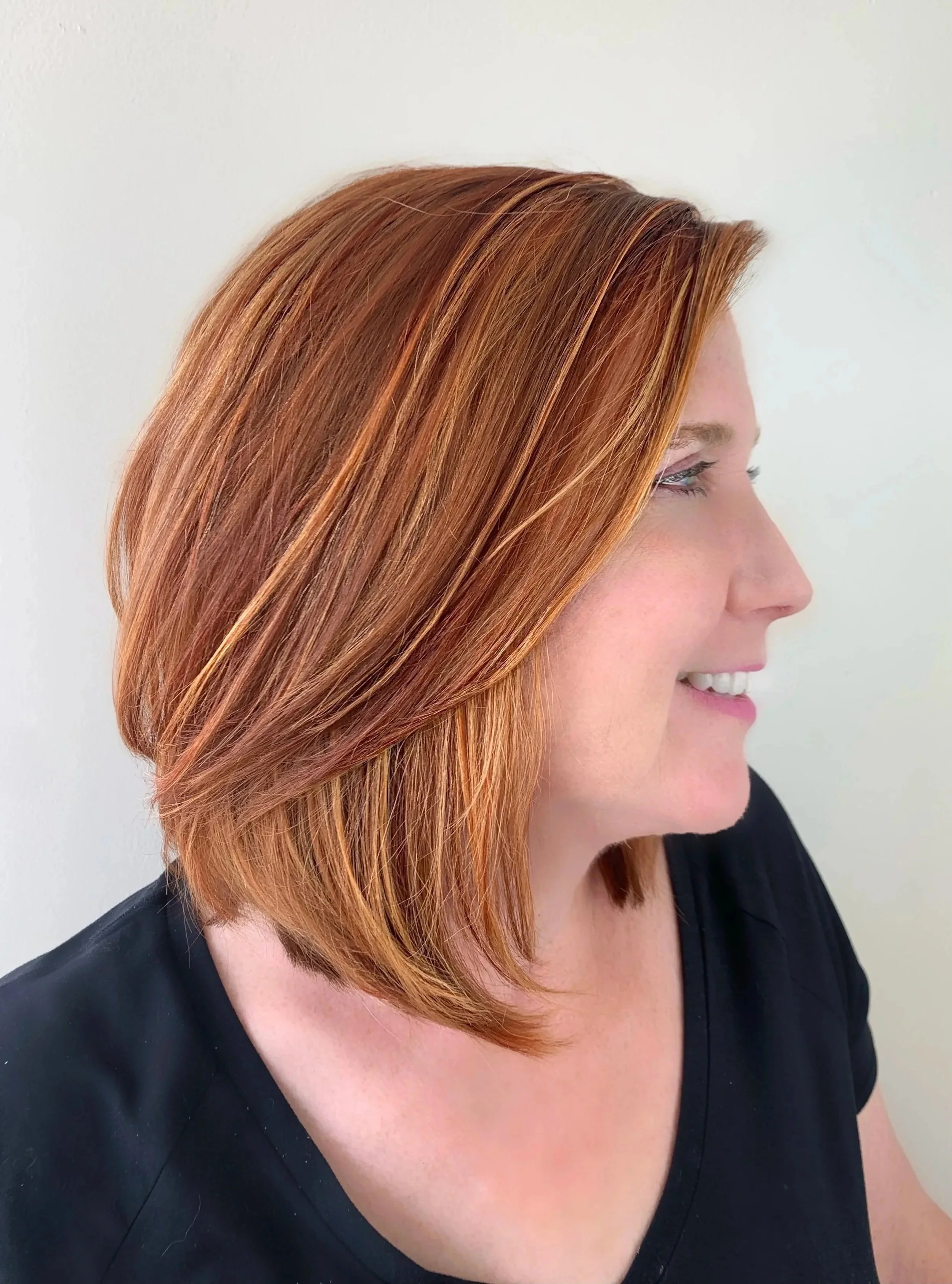 Side profile of a woman with shoulder-length, straight, reddish-brown hair, wearing a black top, against a plain white background.