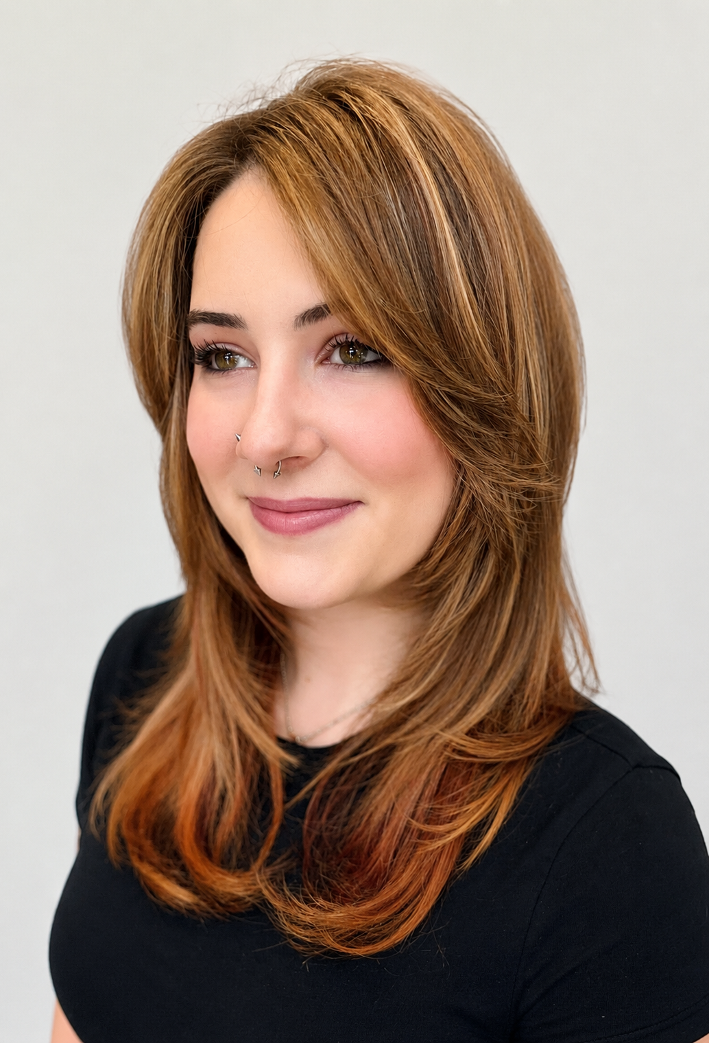A woman with reddish-brown hair styled in layers, wearing a black top, with a septum piercing, smiling softly, and looking slightly to the side against a plain light background.