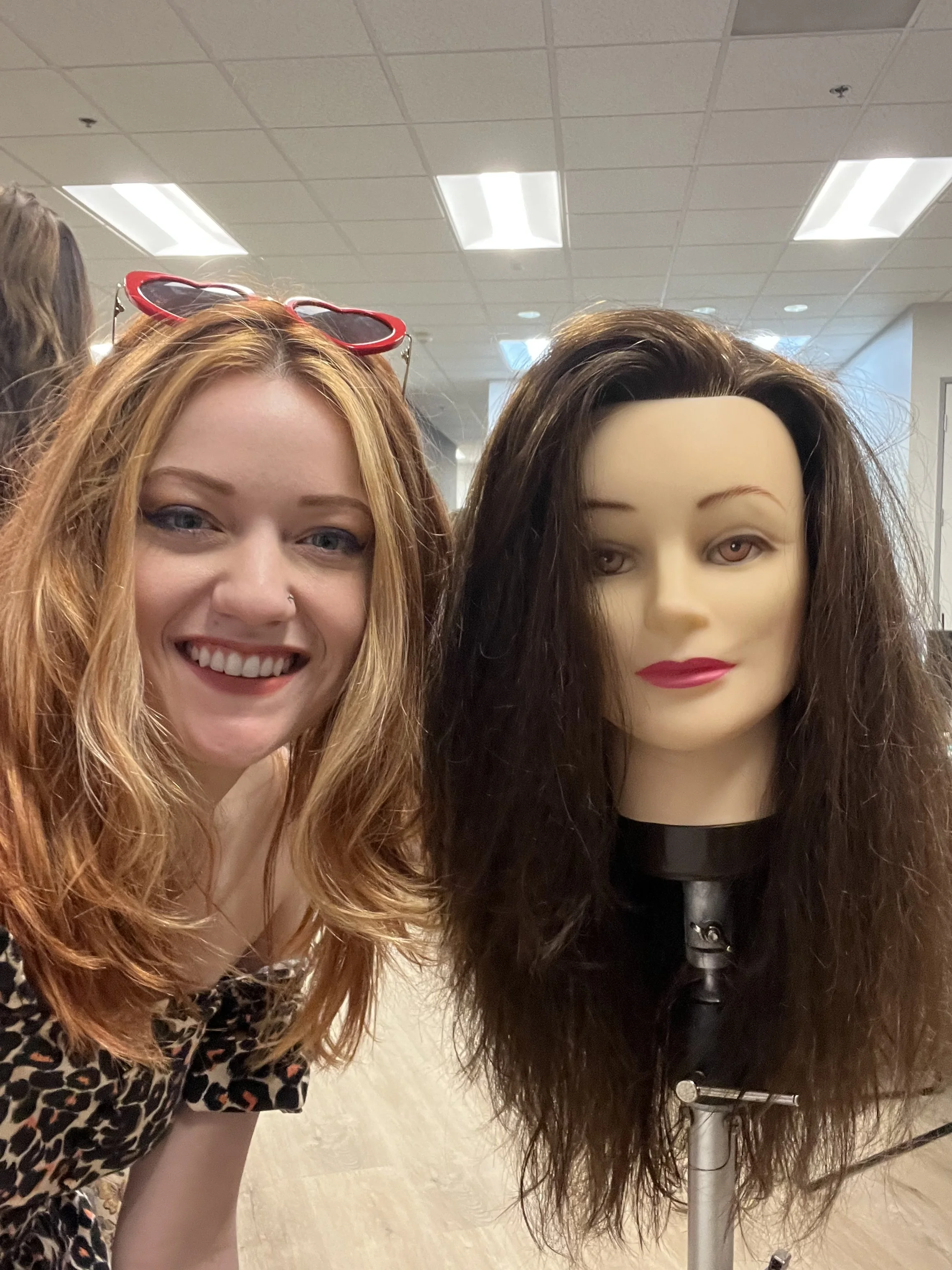 A woman with long, wavy red hair smiling at the camera, wearing red heart-shaped sunglasses on her head, a nose piercing, and a leopard print top. Beside her is a mannequin with long, curly brown hair, wearing red lipstick and a black outfit, standing on a salon or store display stand.