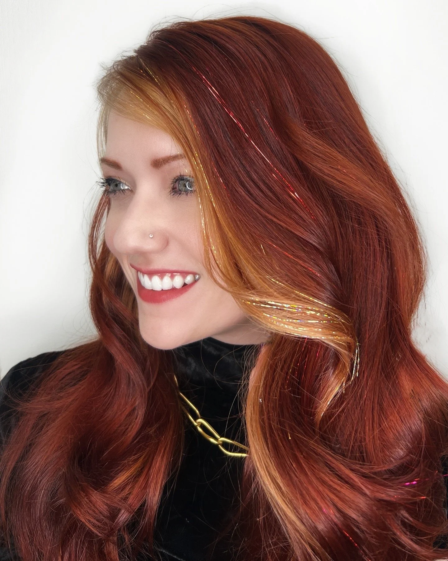 A woman with long, wavy red hair with golden streaks, smiling, wearing a nose stud, a black top, and gold jewelry, against a plain white background.