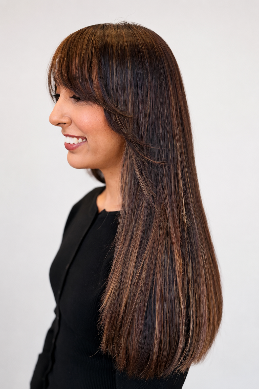 Profile view of a woman with long, straight brown hair, smiling, wearing a black top, against a plain light background.