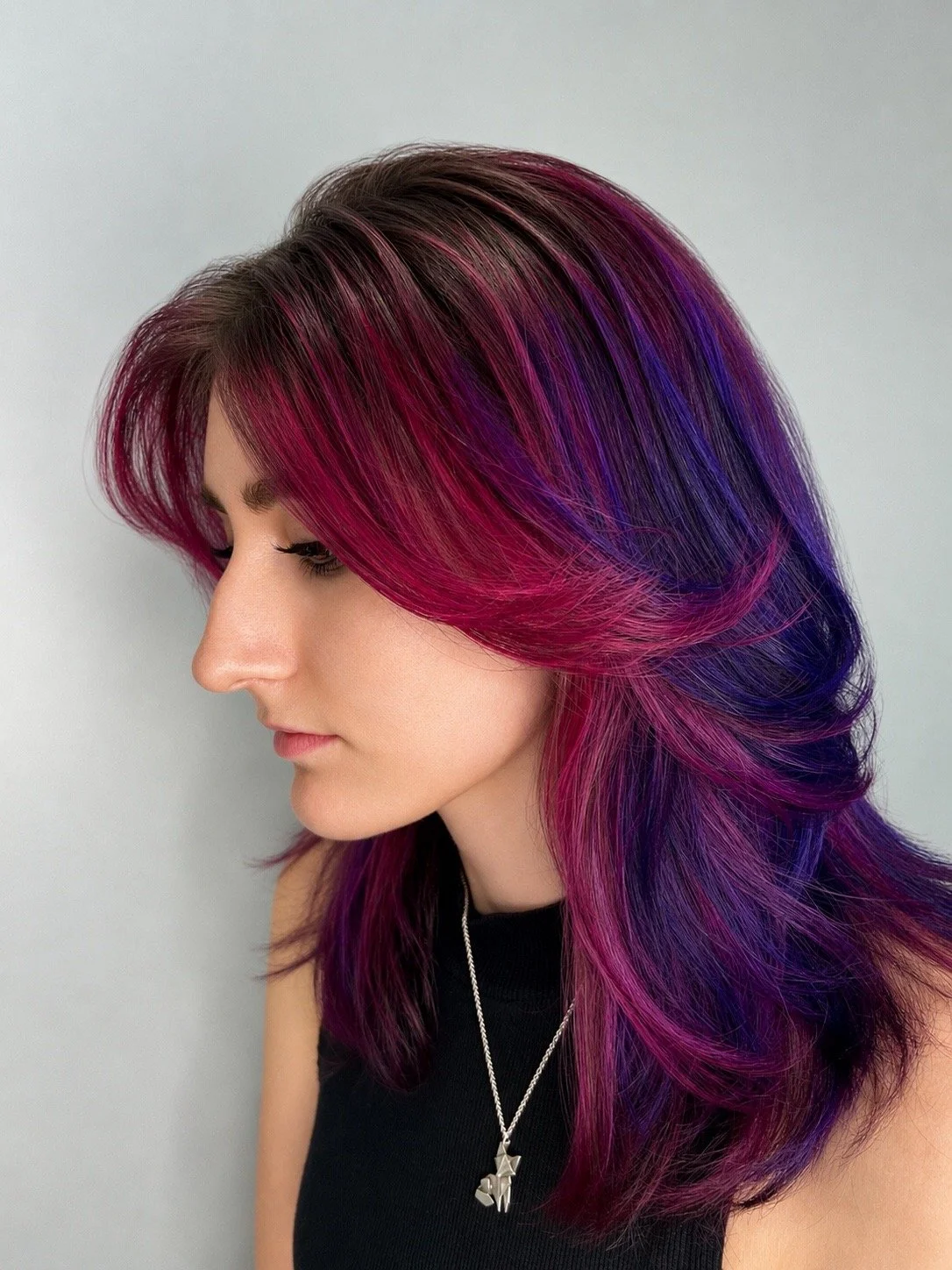 Side profile of a woman with long, layered hair dyed in shades of pink, purple, and blue, wearing a black top and a silver necklace with a geometric pendant.