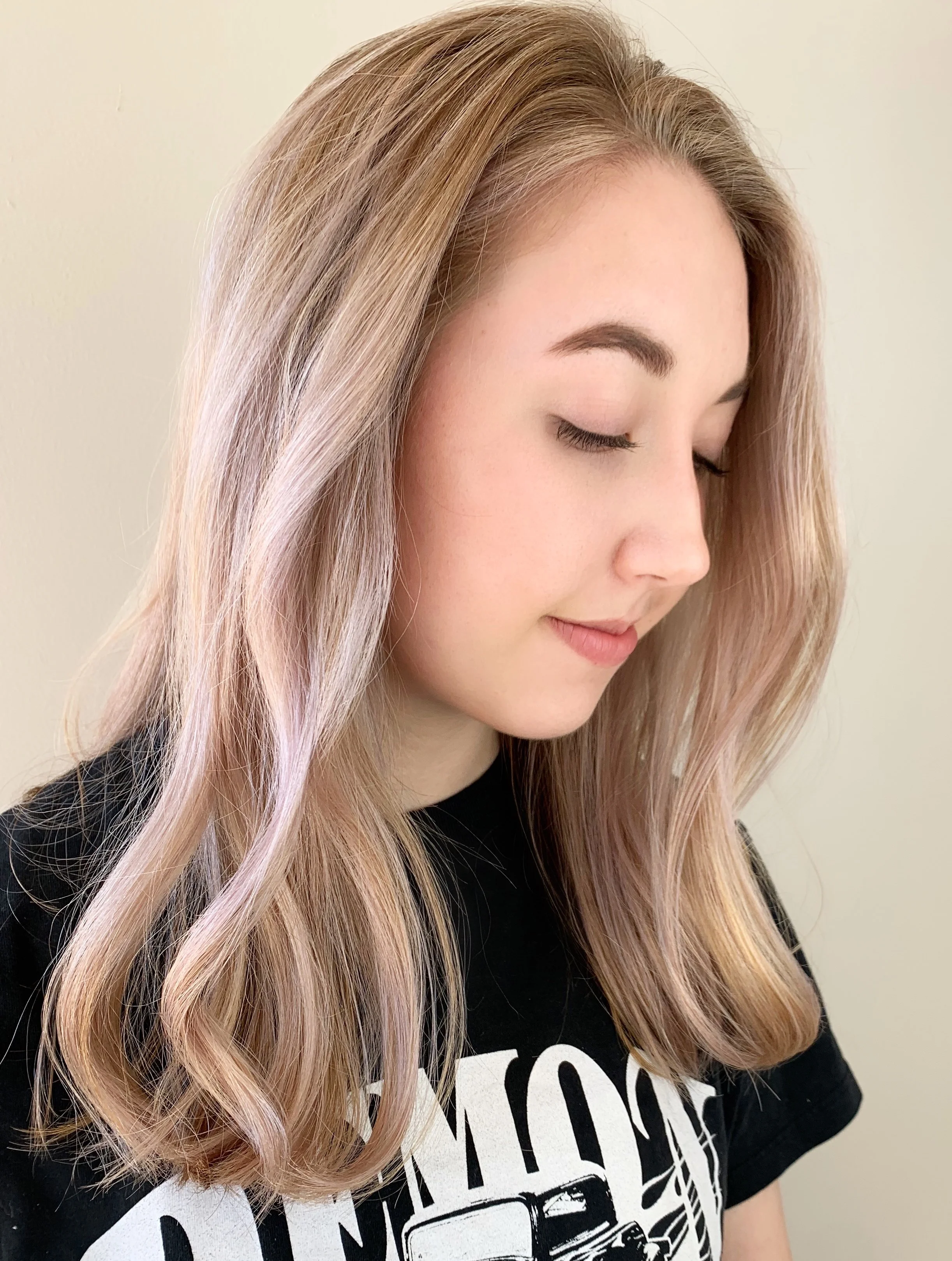 A woman with long, wavy, light-blonde hair with pink shades, looking down with closed eyes, wearing a black t-shirt with a graphic design.