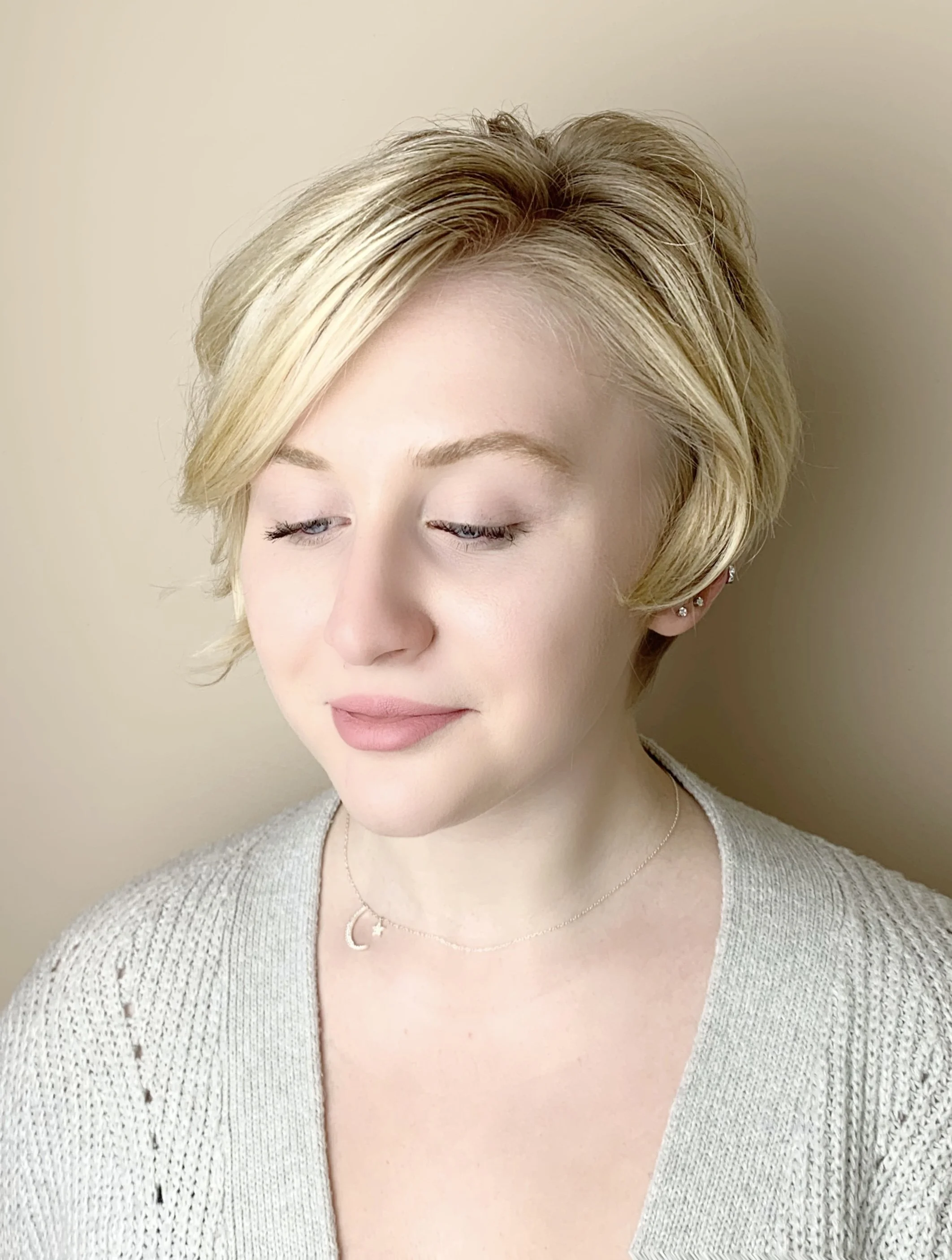Close-up of a woman with short blonde hair, closed eyes, wearing a beige sweater, silver star and moon necklaces, and multiple earrings on her left ear, against a plain wall background.