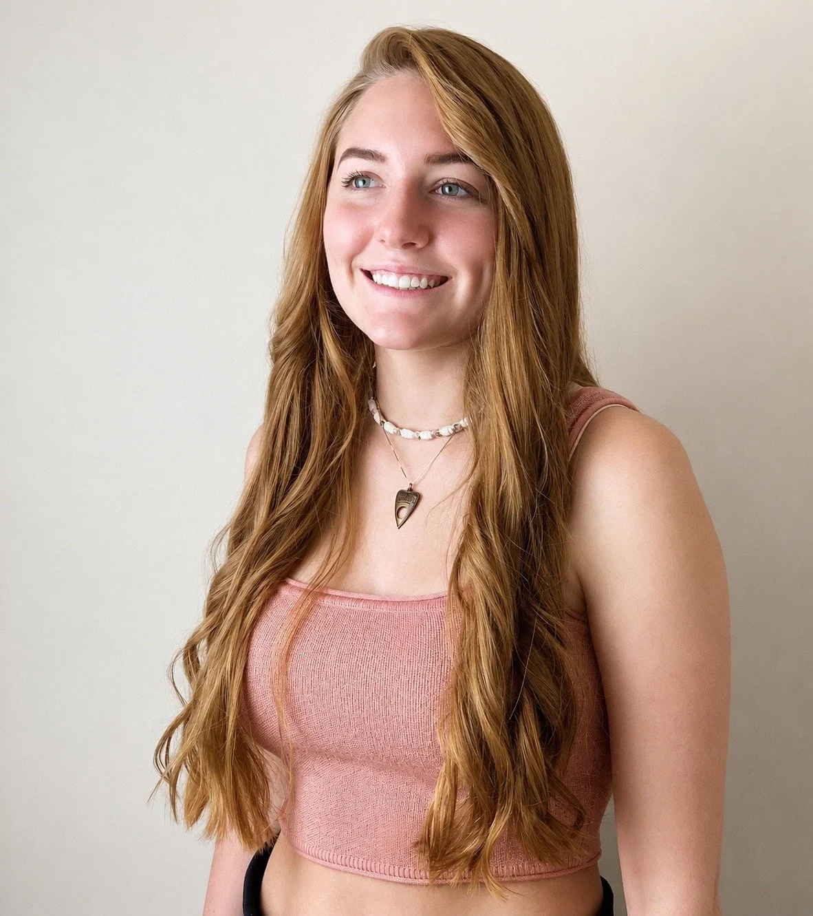 A young woman with long red hair, smiling, wearing a pink tank top and layered necklaces against a plain light-colored background.