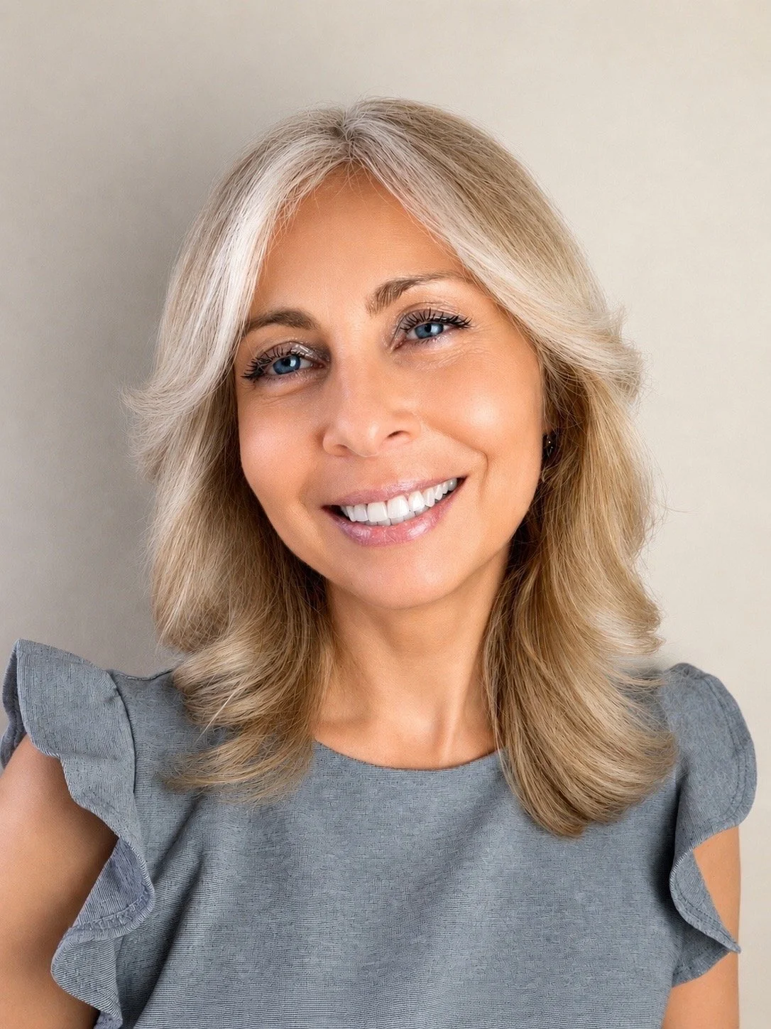 A woman with shoulder-length blonde hair, blue eyes, and a friendly smile, wearing a gray top with ruffled sleeves, standing against a plain light-colored wall.