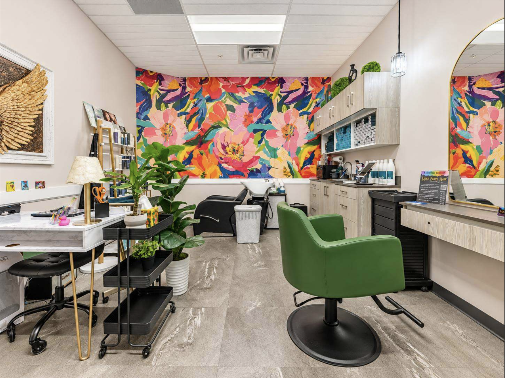 Interior of a colorful hair salon with a vibrant floral mural on the back wall, green salon chairs, a black styling station, and various hair products on shelves.