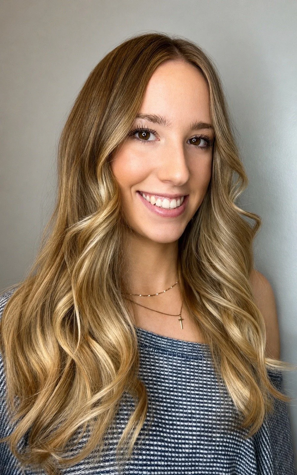 Young woman with long, wavy blonde hair smiling, wearing a blue and white checkered top, a delicate layered necklace with a cross pendant, and standing against a plain gray background.