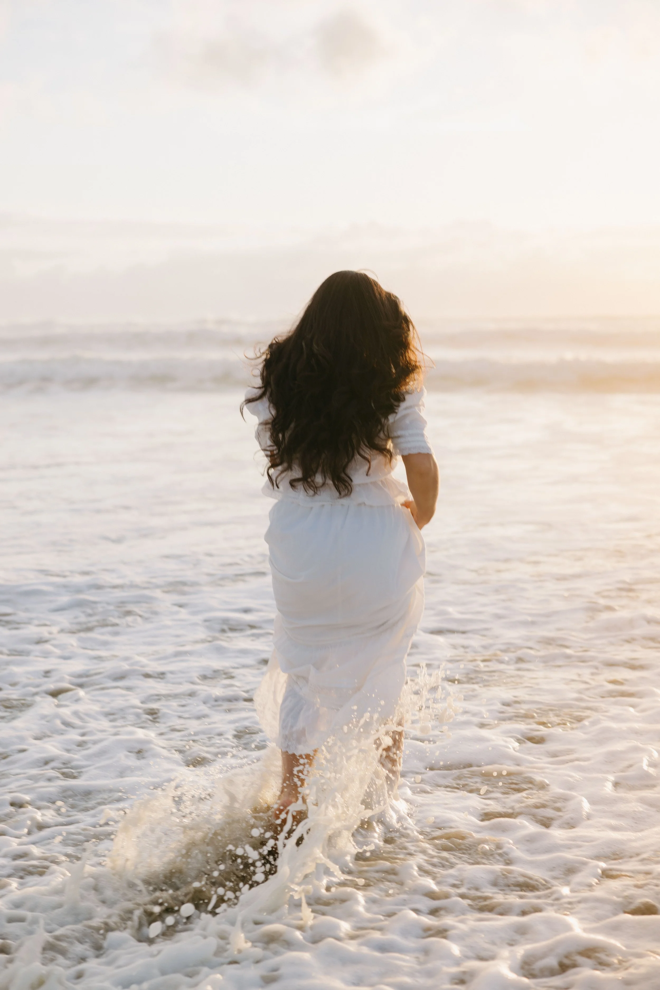 Woman with long dark hair in white dress walking in the ocean at sunset, splashing water as she moves.