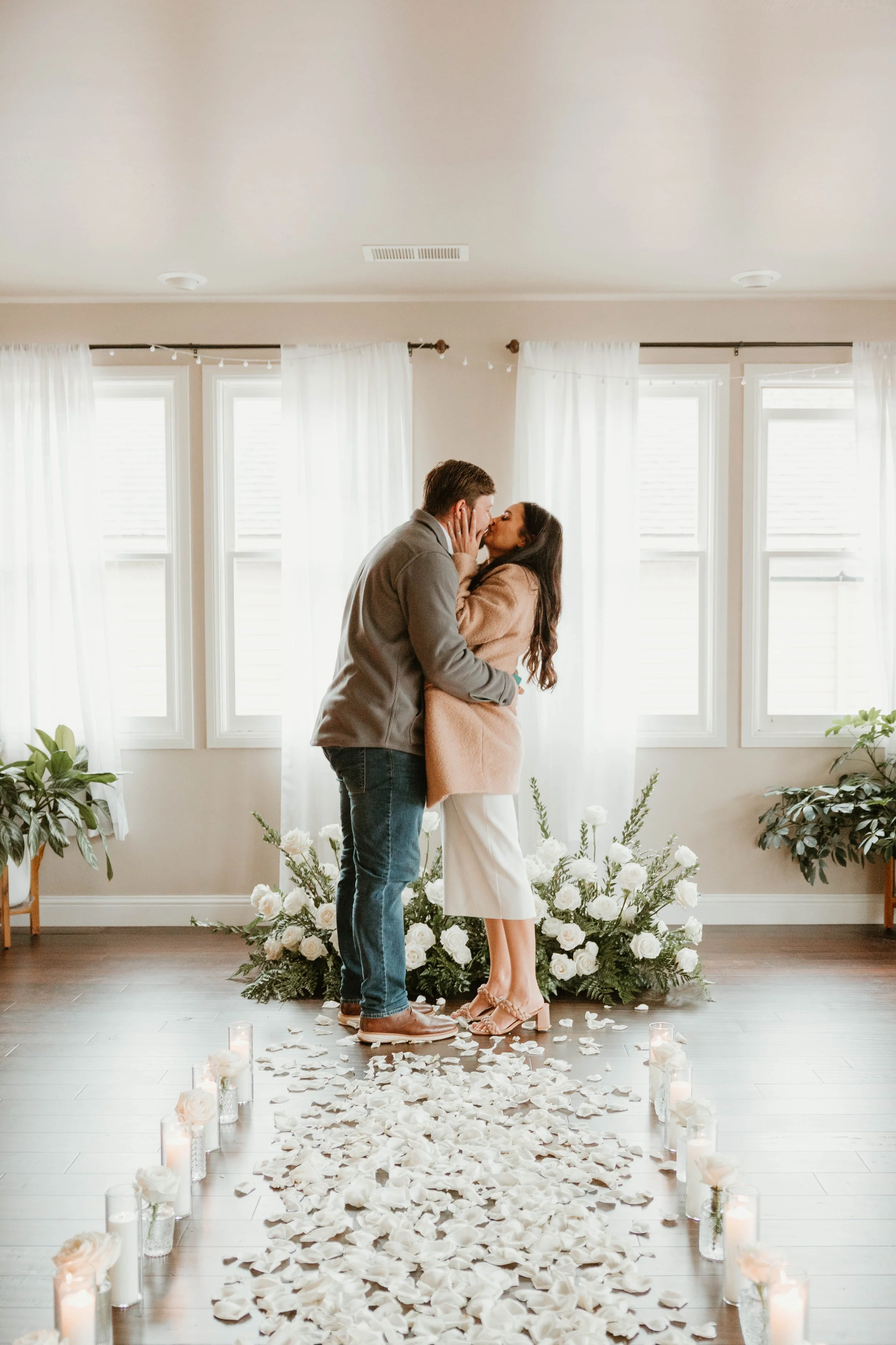 A couple is sharing a kiss in a room decorated with white flowers and candles, with white curtains and multiple windows in the background. The floor is strewn with white petals.
