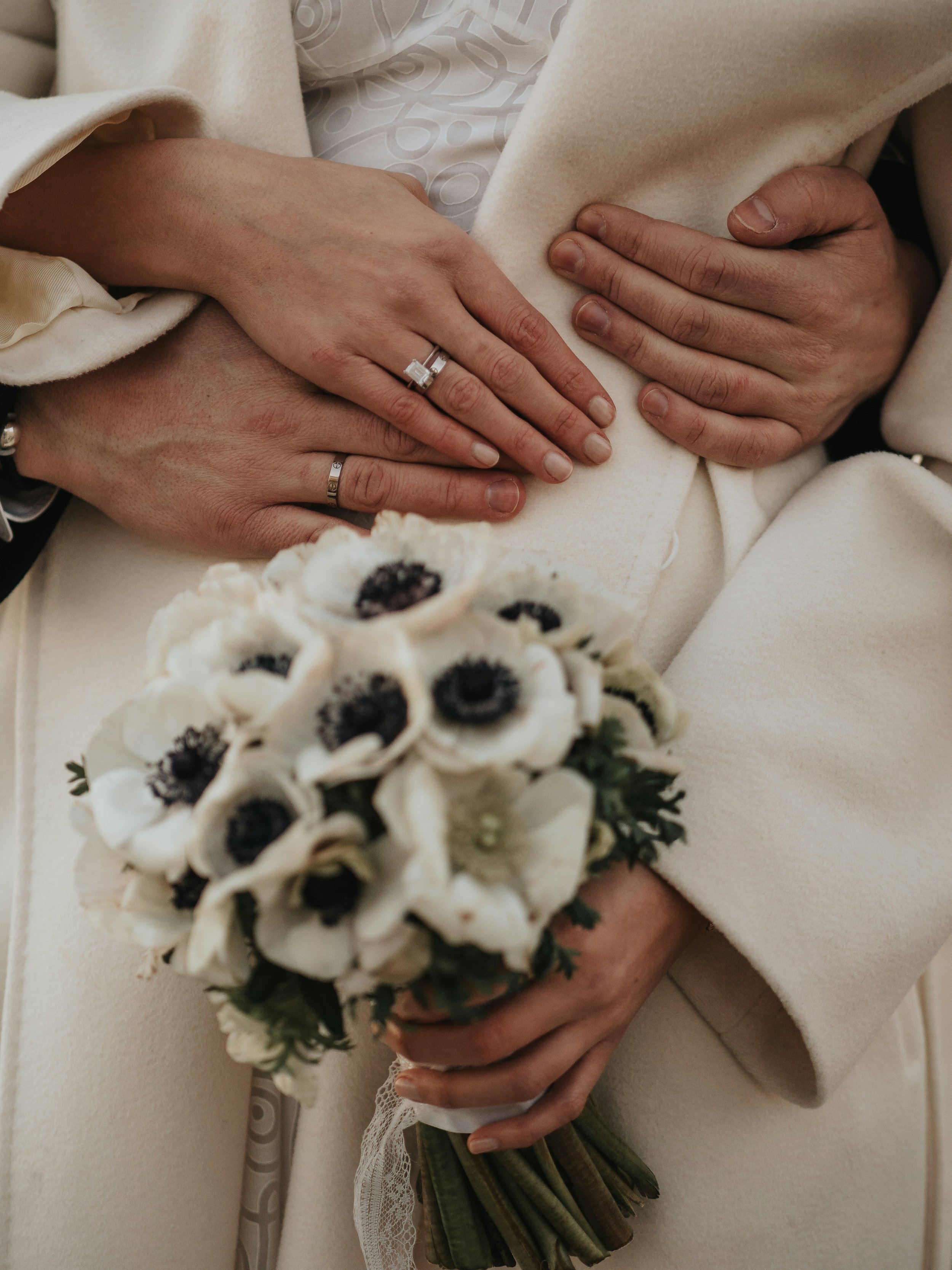 Close-up of two hands holding a bouquet of white flowers with dark centers, wearing wedding rings, and dressed in formal clothing.