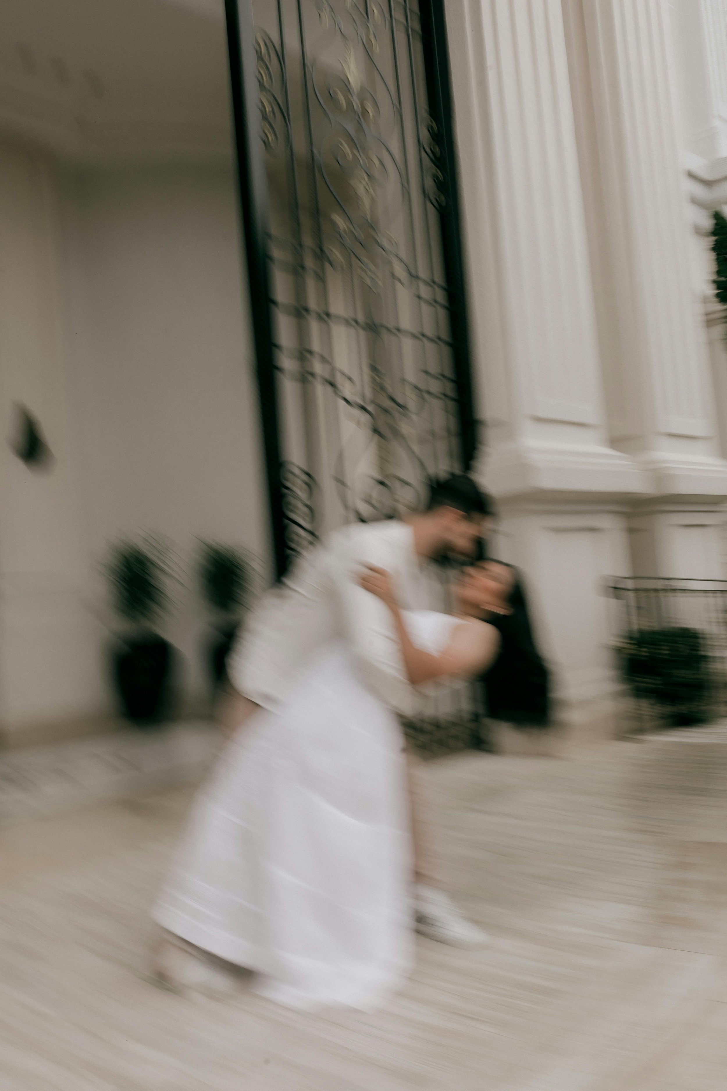 A blurred photo of two women in white wedding dresses, standing indoors with elegant decor, including a wrought iron railing and white draped curtains.