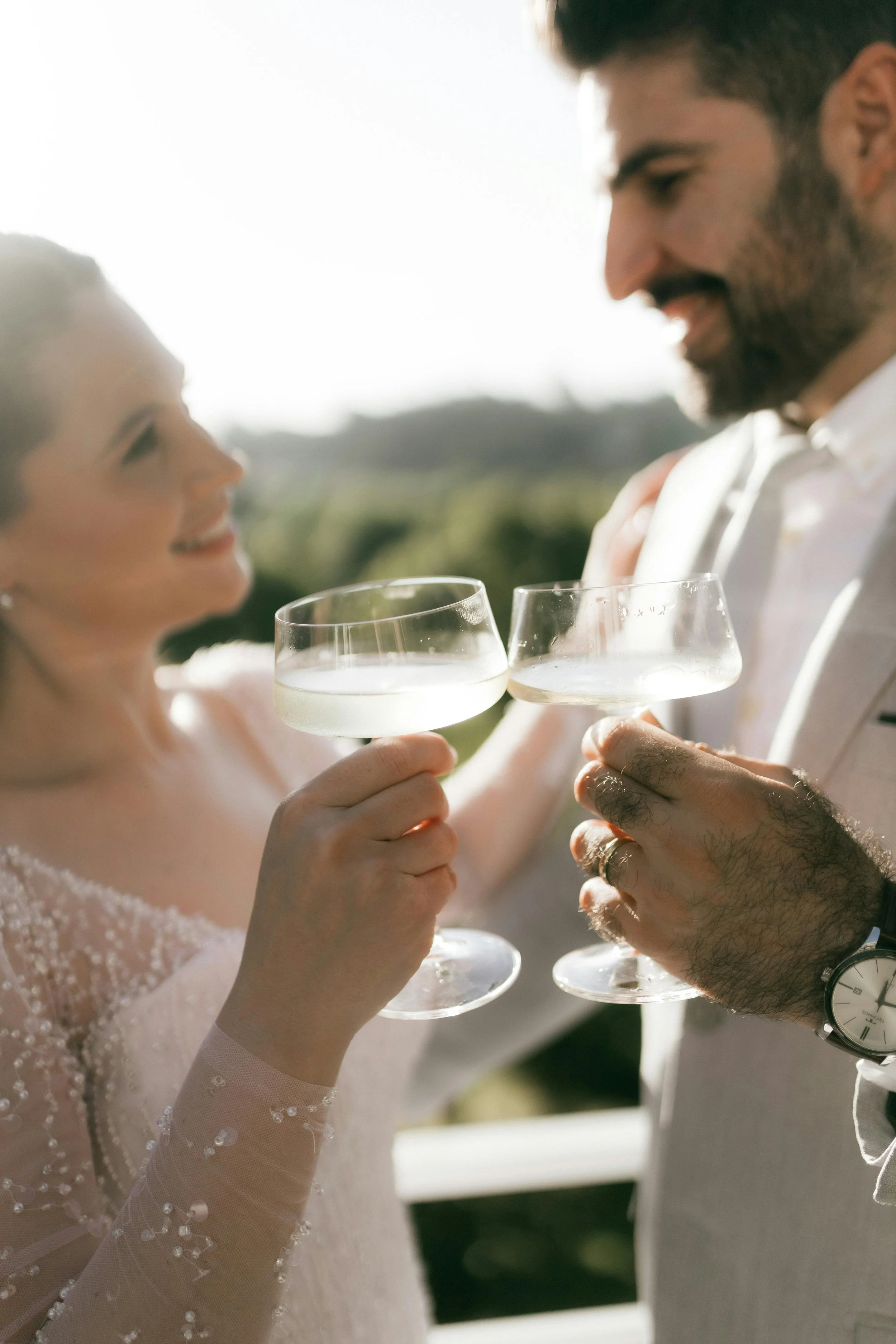 A couple smiling and holding glasses of champagne, likely celebrating an event, outdoors with bright sunlight.
