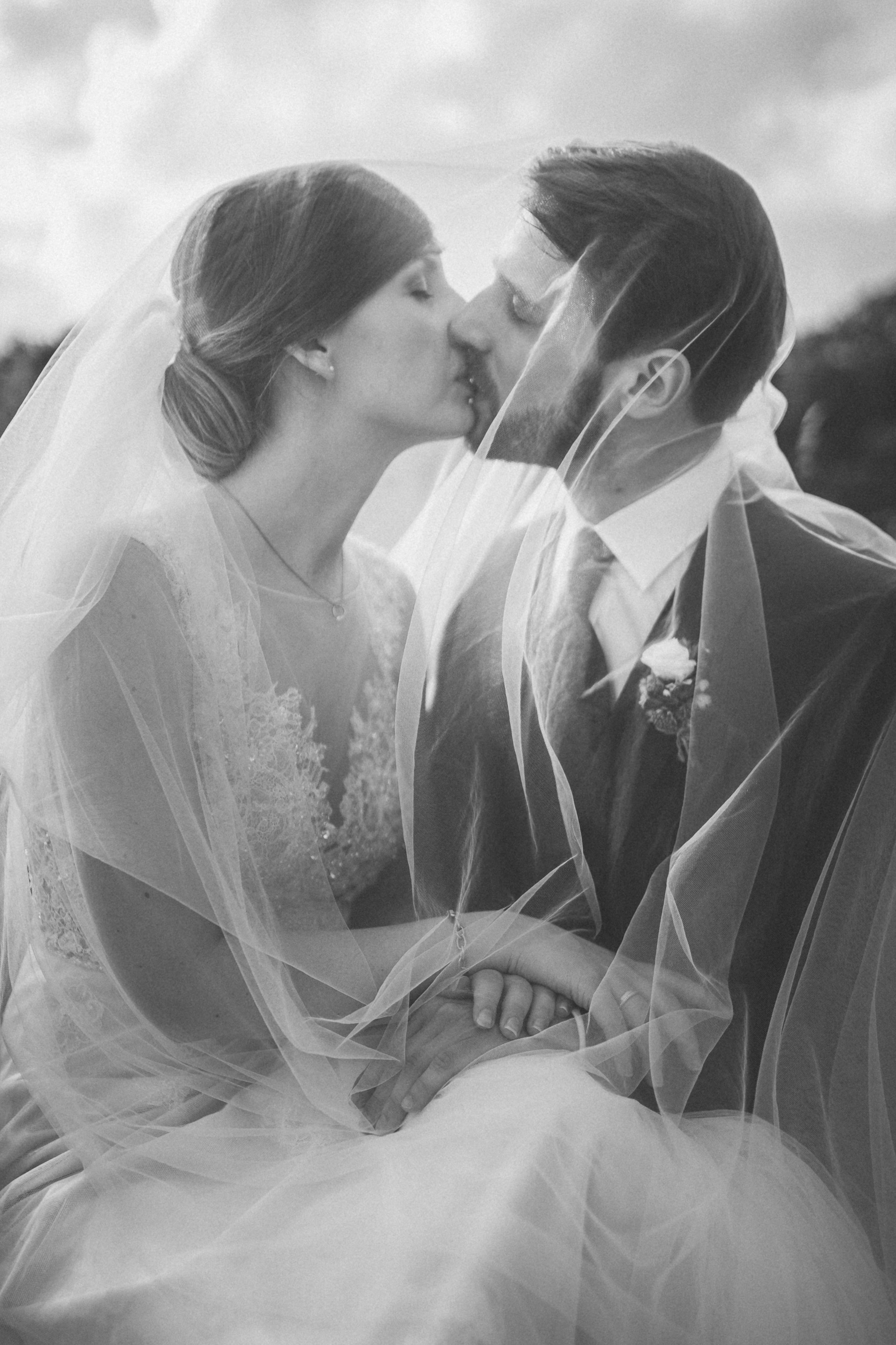 Black and white photo of a bride and groom sharing a kiss, with veil and wedding attire, outside during daytime.