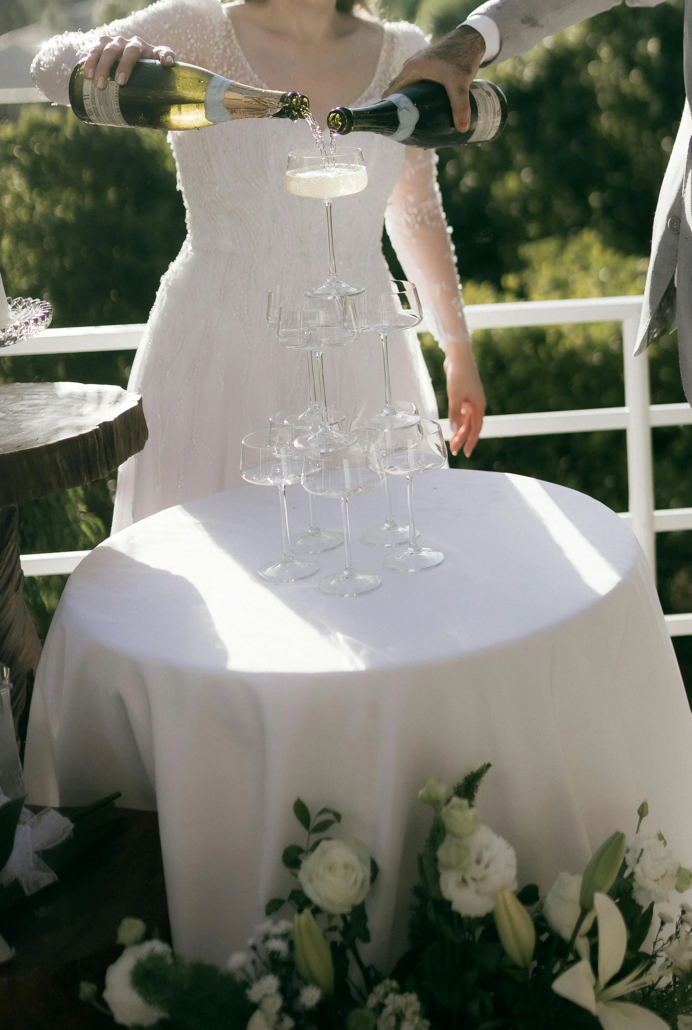 People pouring champagne into a glass at a wedding reception, with a tiered champagne glass stand, flowers, and white drapery in the background.