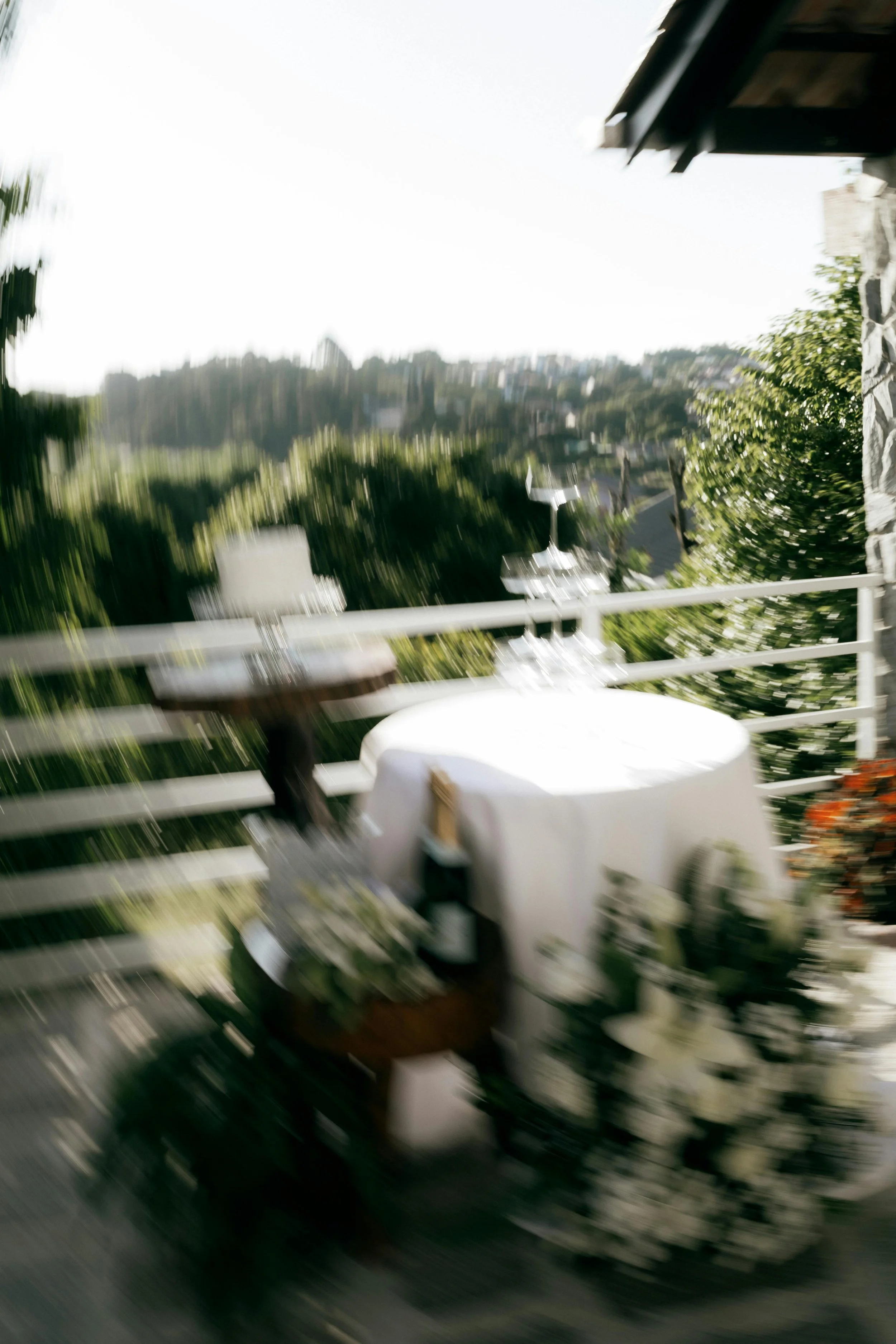 Blurred image of an outdoor patio or balcony with a white railing, some greenery, and an outdoor table, with a distant view of a hillside with houses.