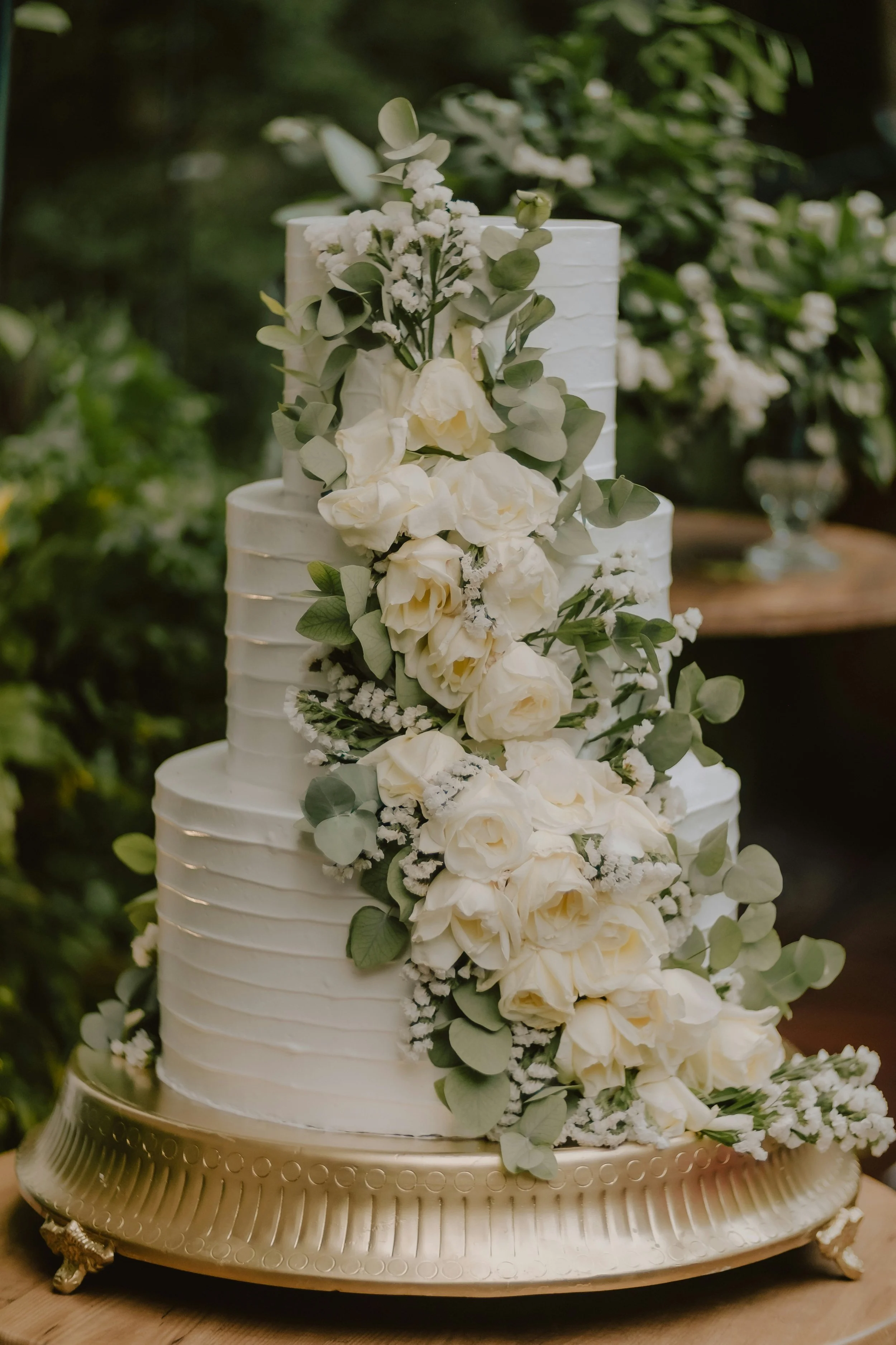A tall white wedding cake decorated with white roses, eucalyptus leaves, and small white flowers, placed on a gold cake stand.