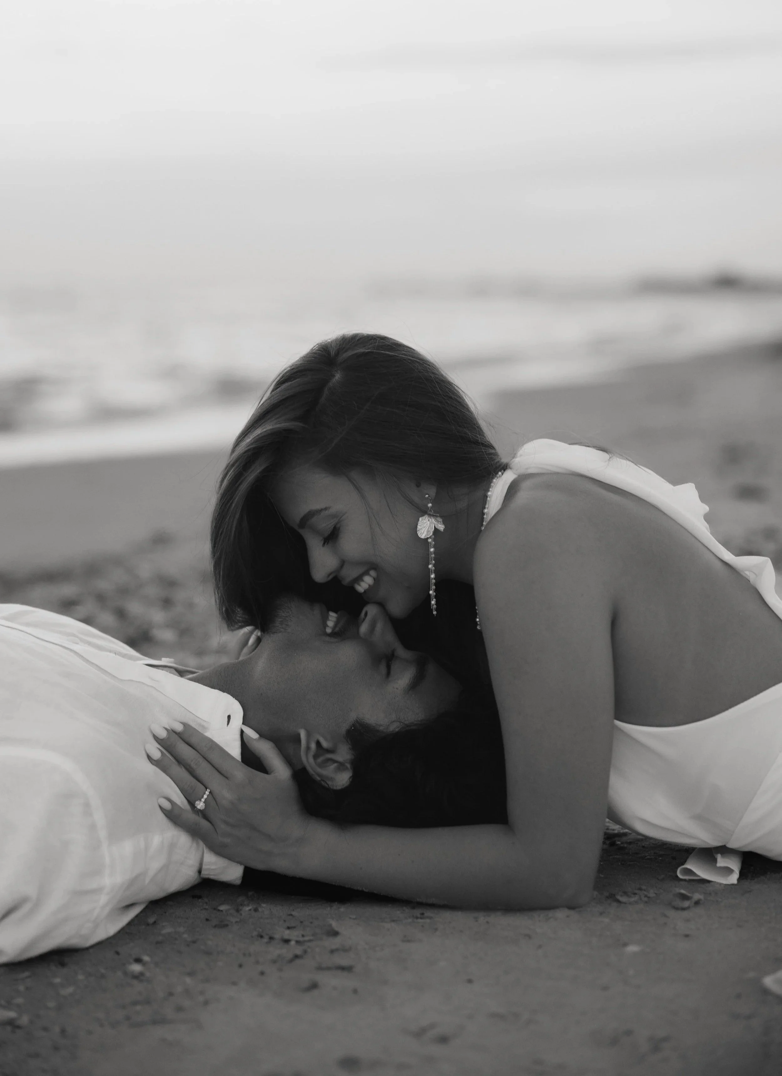A black and white photo of two women on a beach, smiling and touching foreheads, with one woman lying on the sand and the other leaning over her.