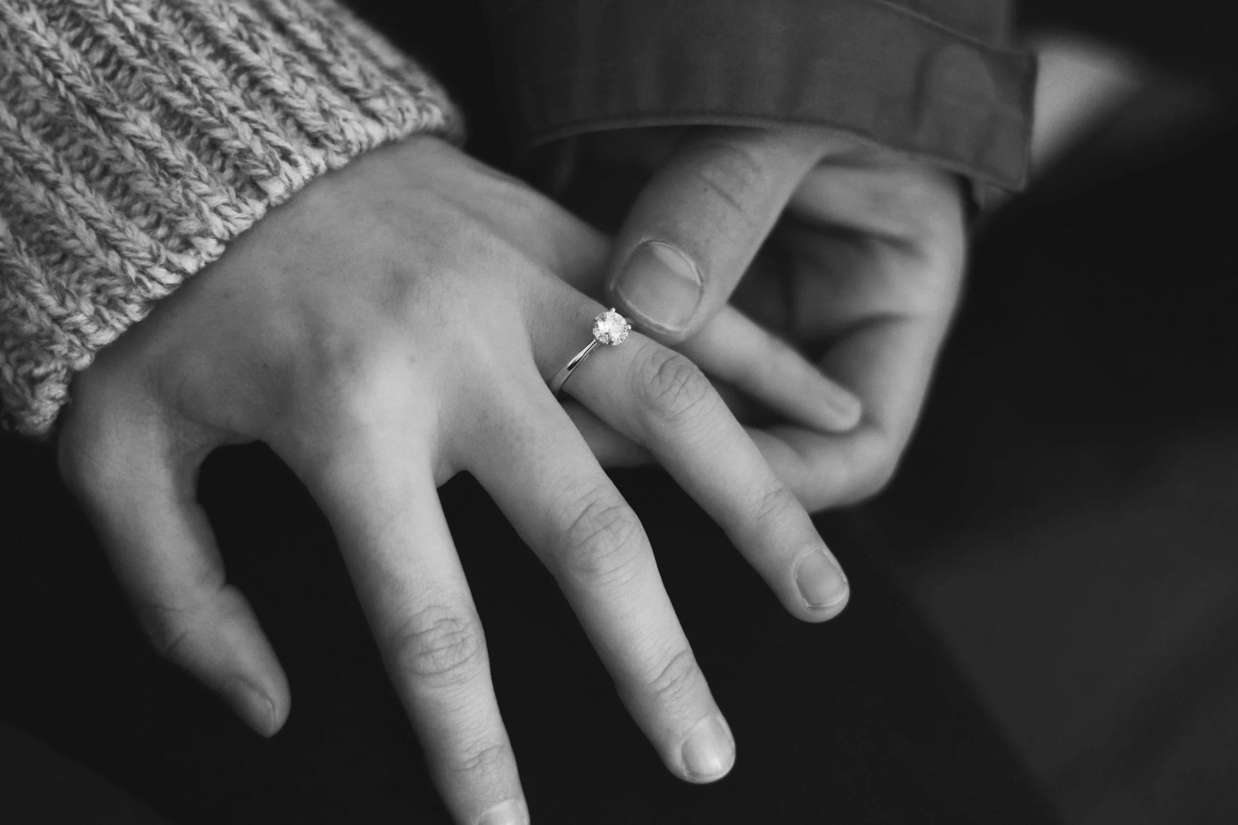 Close-up of a hand wearing a ring with a large gemstone, with another hand gently touching the ring, on a dark background.
