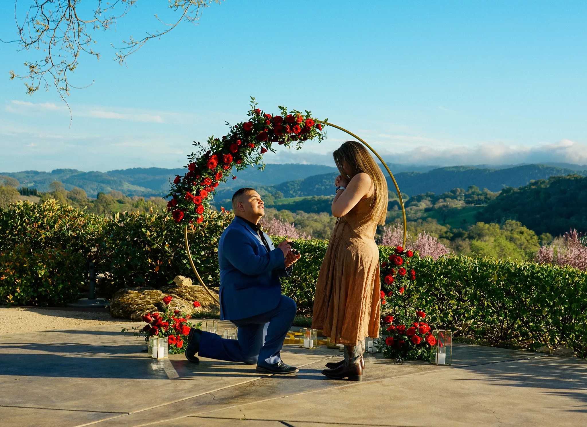 A man kneeling and proposing marriage to a woman on a scenic outdoor location with mountains, trees, and clear blue sky in the background. The woman is standing with her hands covering her face, and the man is holding a small box, likely an engagement ring. There is a decorative arch adorned with red flowers behind them, and candles and flowers are arranged on the ground around them.