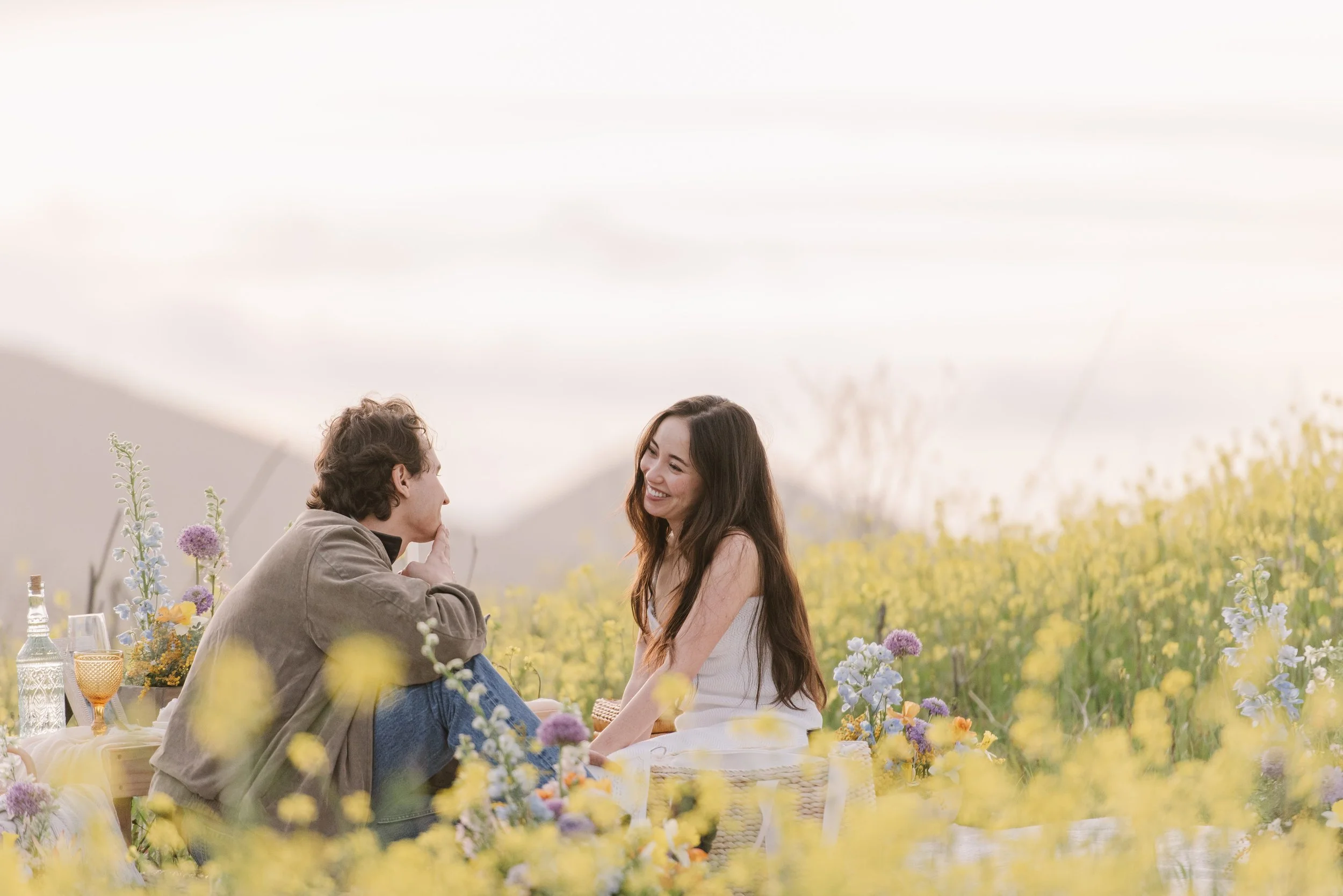 A young man and woman are enjoying a romantic moment at an outdoor wedding reception, surrounded by flowers and candles on a table, with a scenic mountain backdrop.