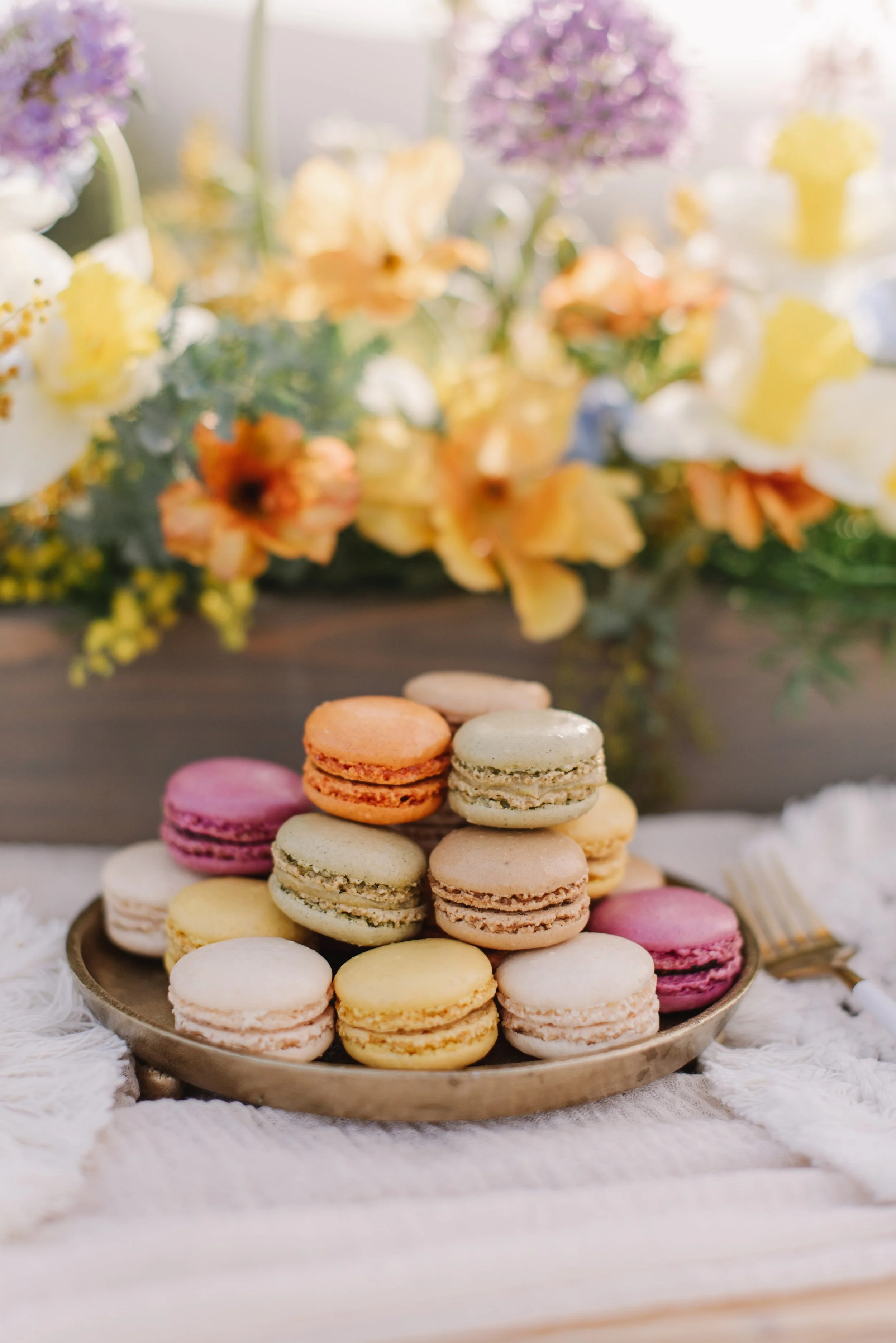 Stacked French macarons in pastel colors on a silver tray with a blurred floral background.