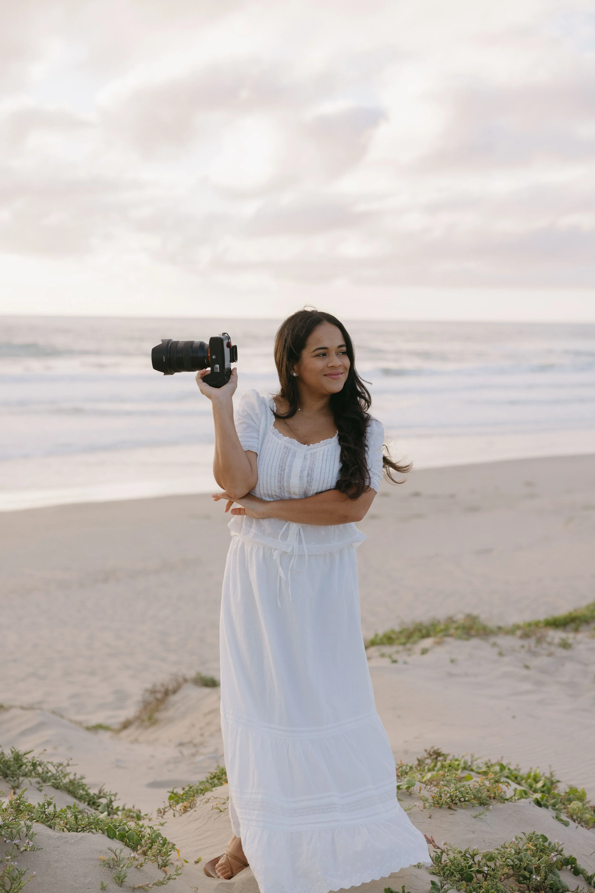 A woman standing on a sandy beach holding a camera in her right hand and smiling. She is wearing a white dress and sandals, with the ocean and cloudy sky in the background.