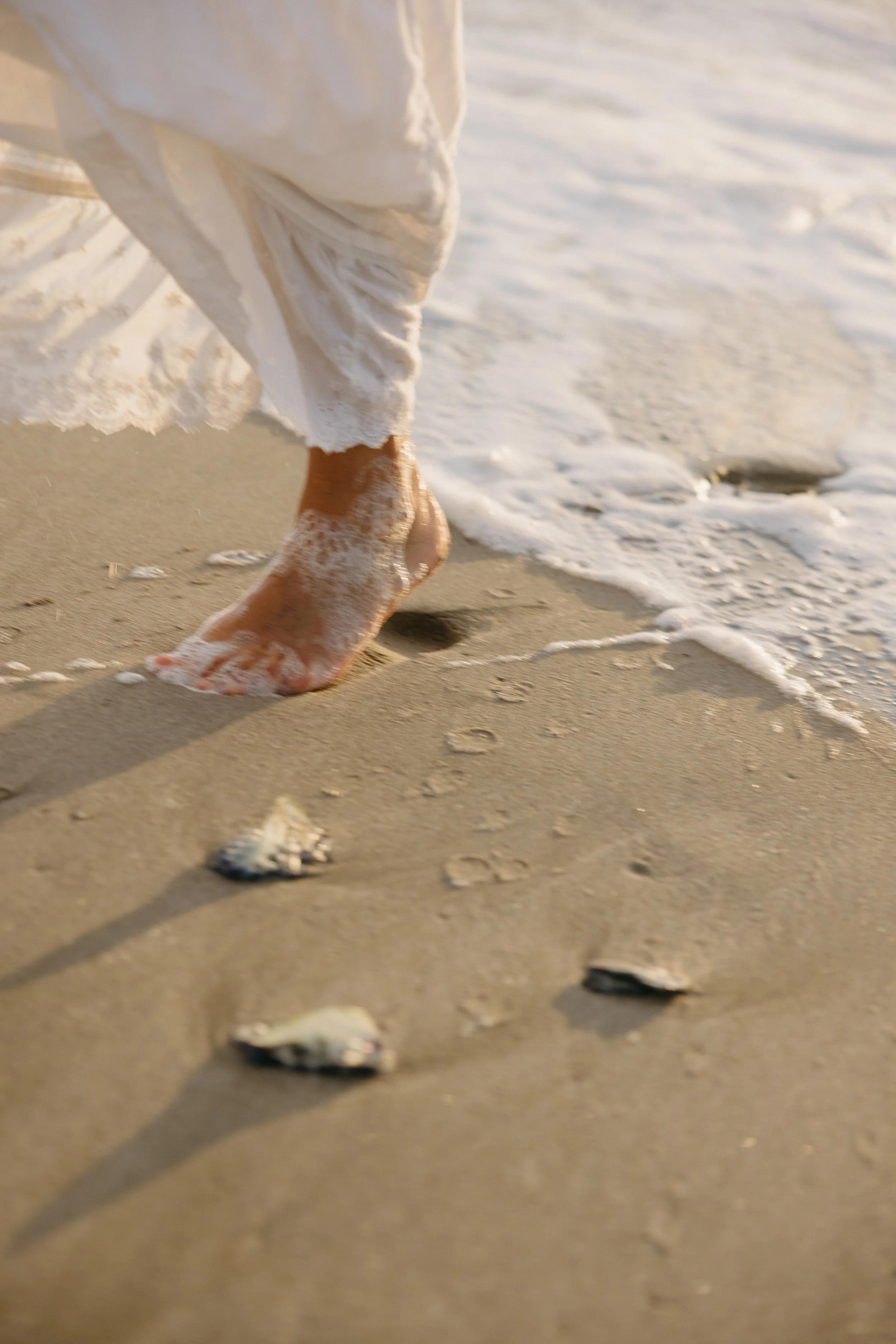 Person walking along the beach with bare feet, white dress, and ocean waves nearby.