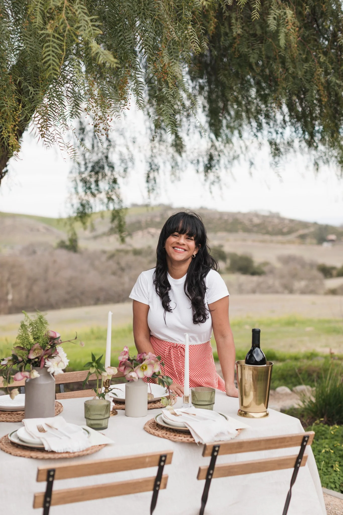 A smiling woman with dark hair and bangs wearing a white top and red striped apron standing outdoors at a decorated table with flowers, candles, and wine, with a rural landscape and trees in the background.