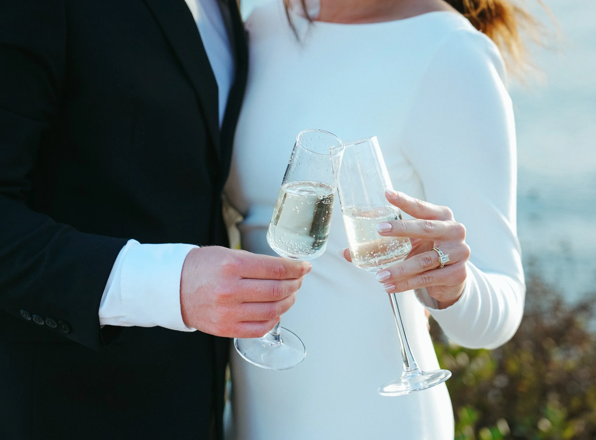 Two people, dressed in formal attire, are holding champagne glasses and toasting. The background appears to be outdoors with natural lighting.