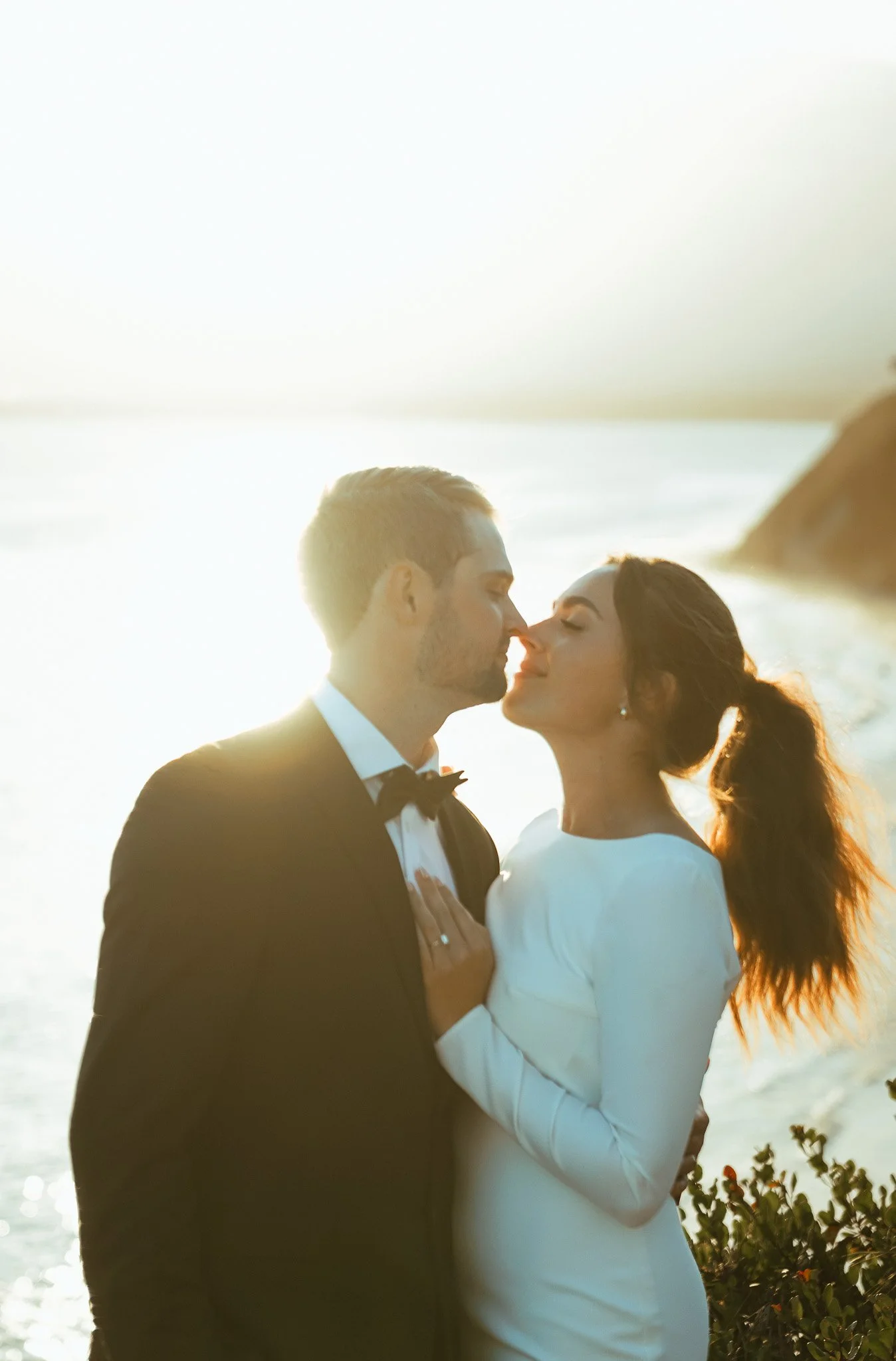 A newlywed couple stands close to each other at the beach during what appears to be a sunset, with their noses touching and eyes closed, dressed in wedding attire, backlit by the bright sun creating a warm glow.