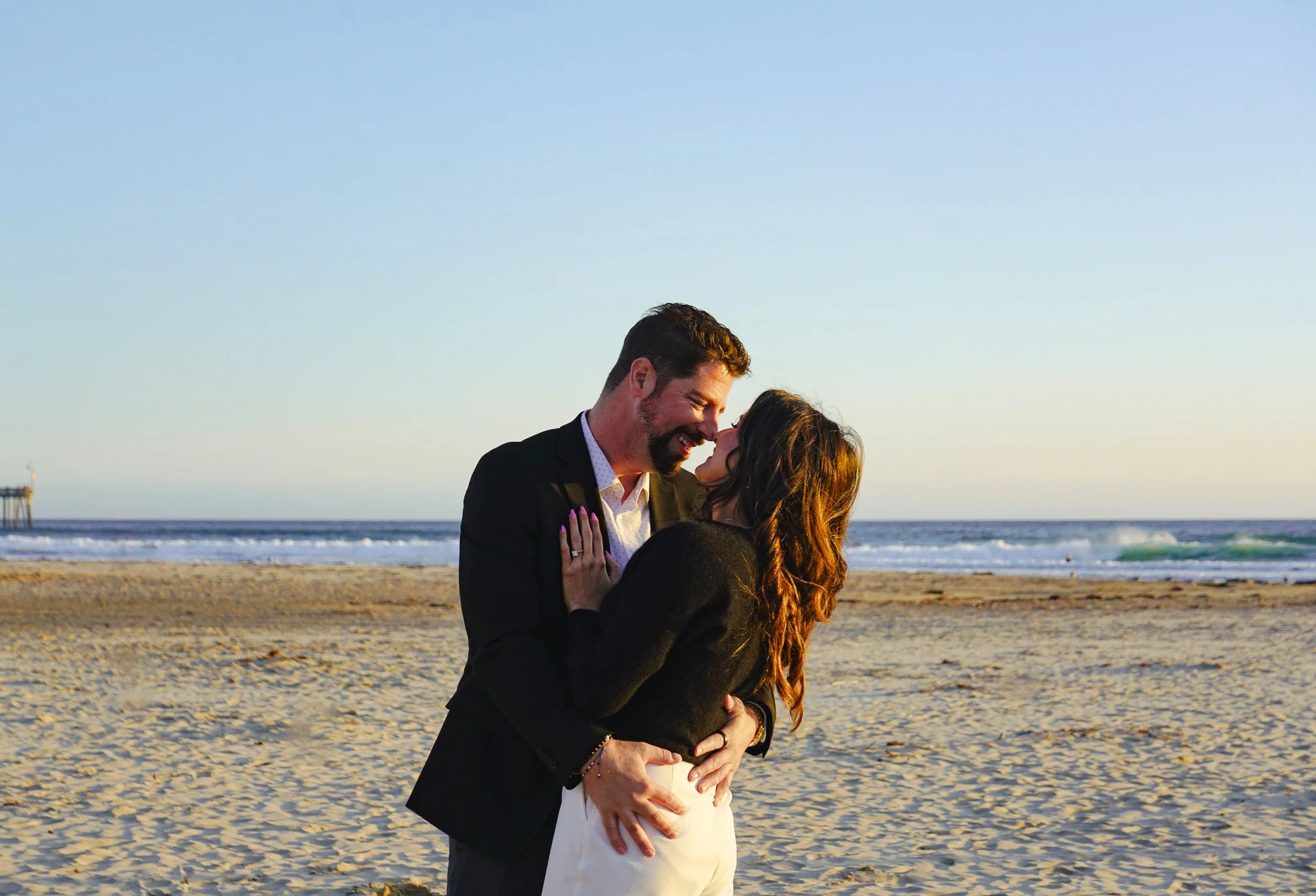 A couple embracing and smiling at each other on a beach with the ocean in the background during sunset.