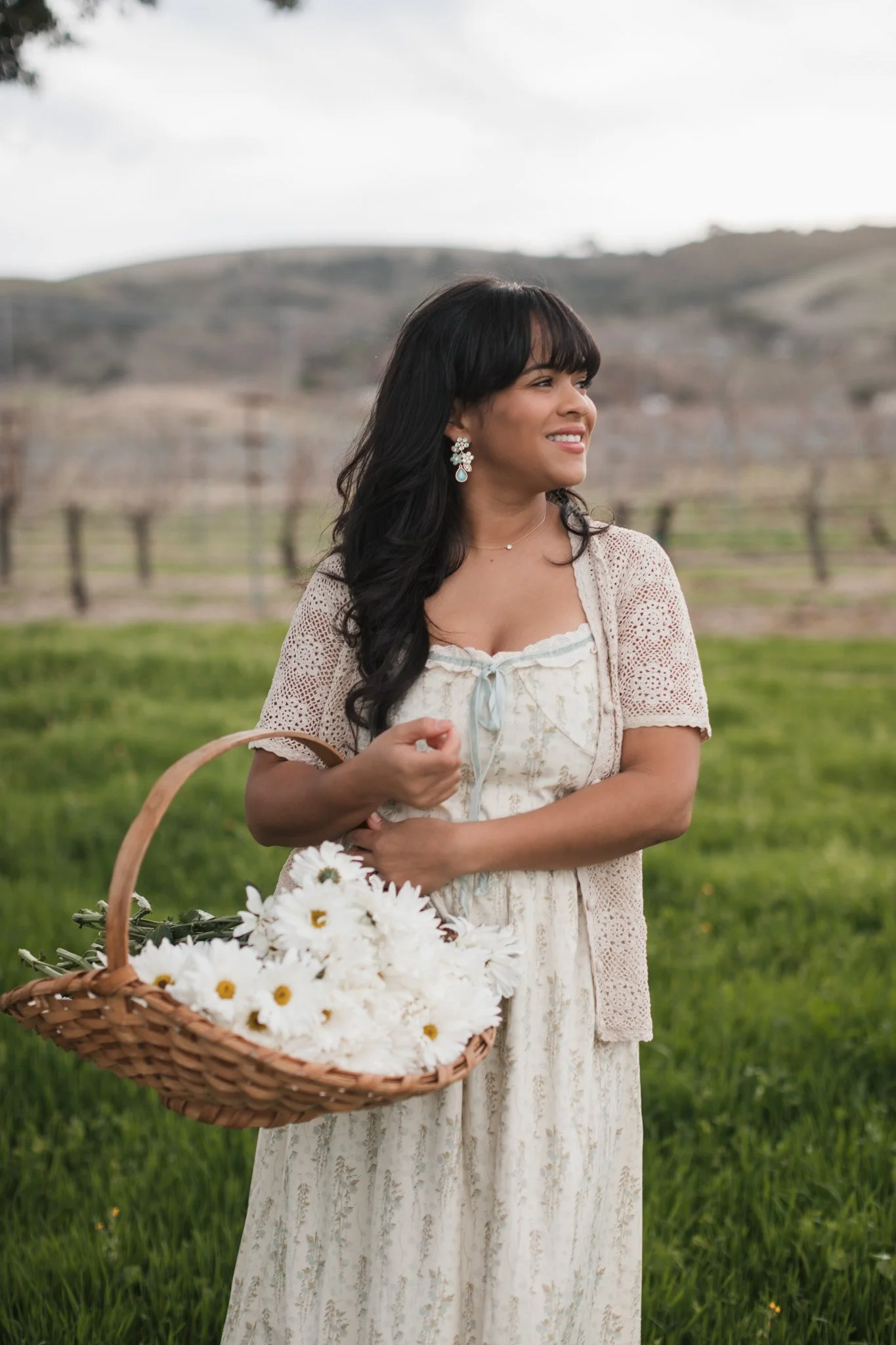 A woman standing outdoors in a grassy field, holding a wicker basket filled with white daisies, with a rural landscape of hills and a fence in the background.