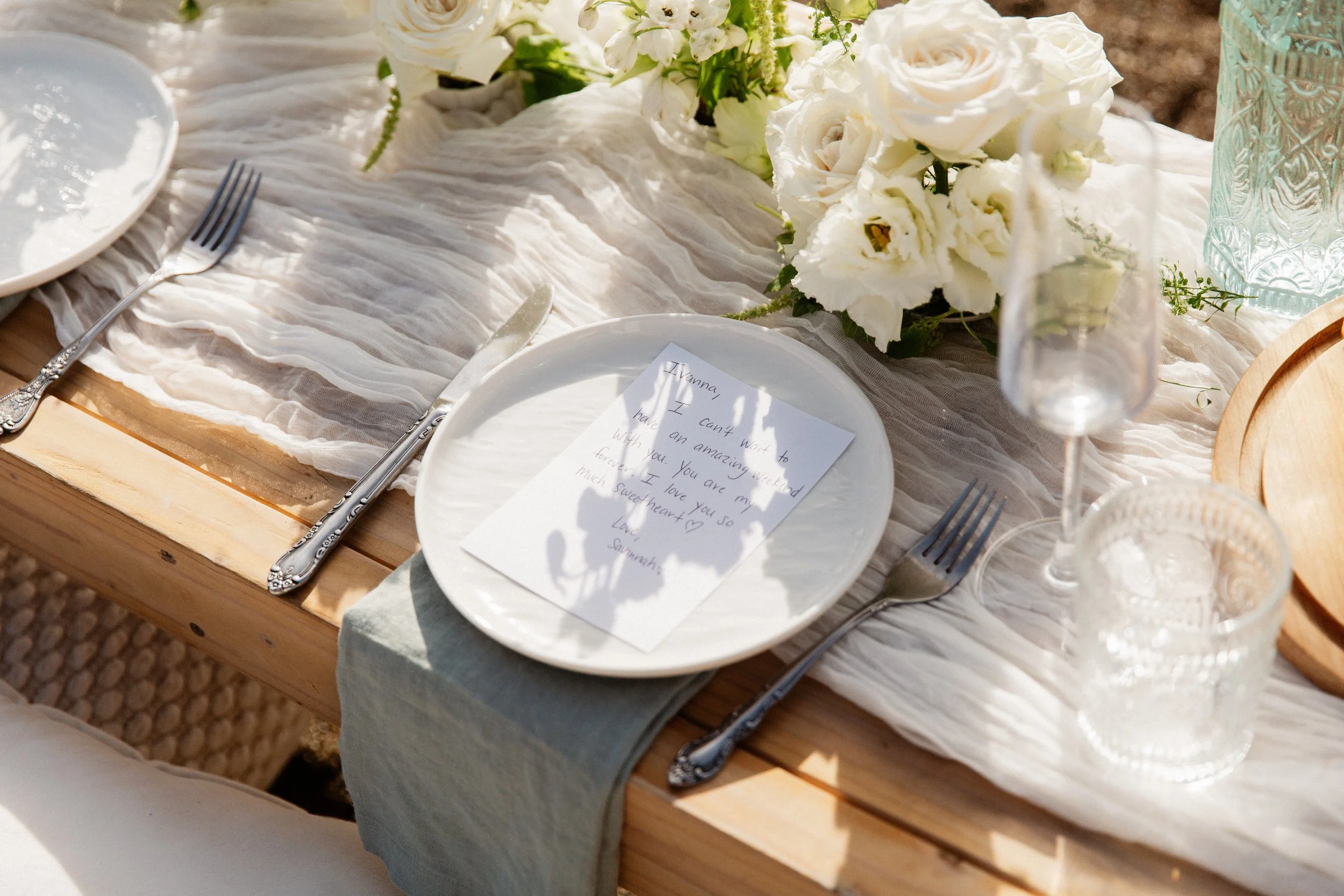 A table set for an event or celebration with a centerpiece of white flowers, including roses and other blooms, on a white fabric tablecloth. There are plates, silverware, a handwritten note, and decorative glassware on the table, suggesting a formal or special occasion like a wedding or brunch.