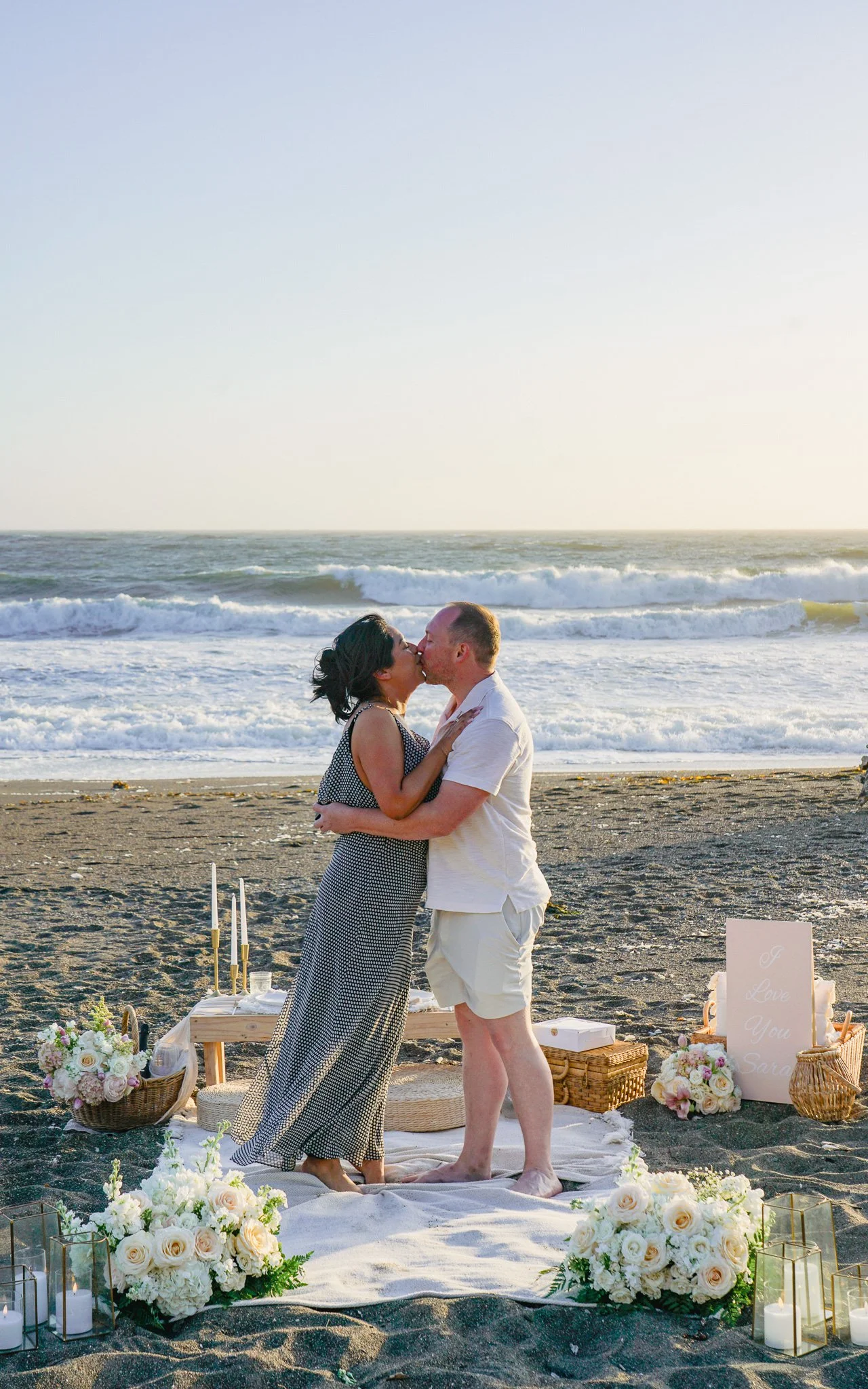 A couple is kissing during a wedding ceremony on a beach, with ocean waves in the background. They are standing on a decorated area with flowers, candles, and a small sign that reads 'Love You Sara'.