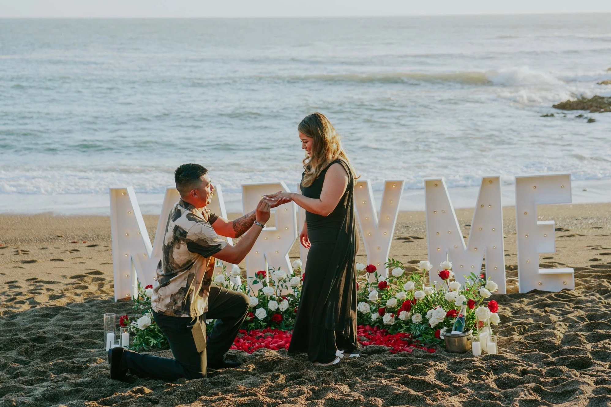 A couple getting engaged on a beach with a romantic floral display, candles, and large illuminated letters spelling 'LOVE' in the background.