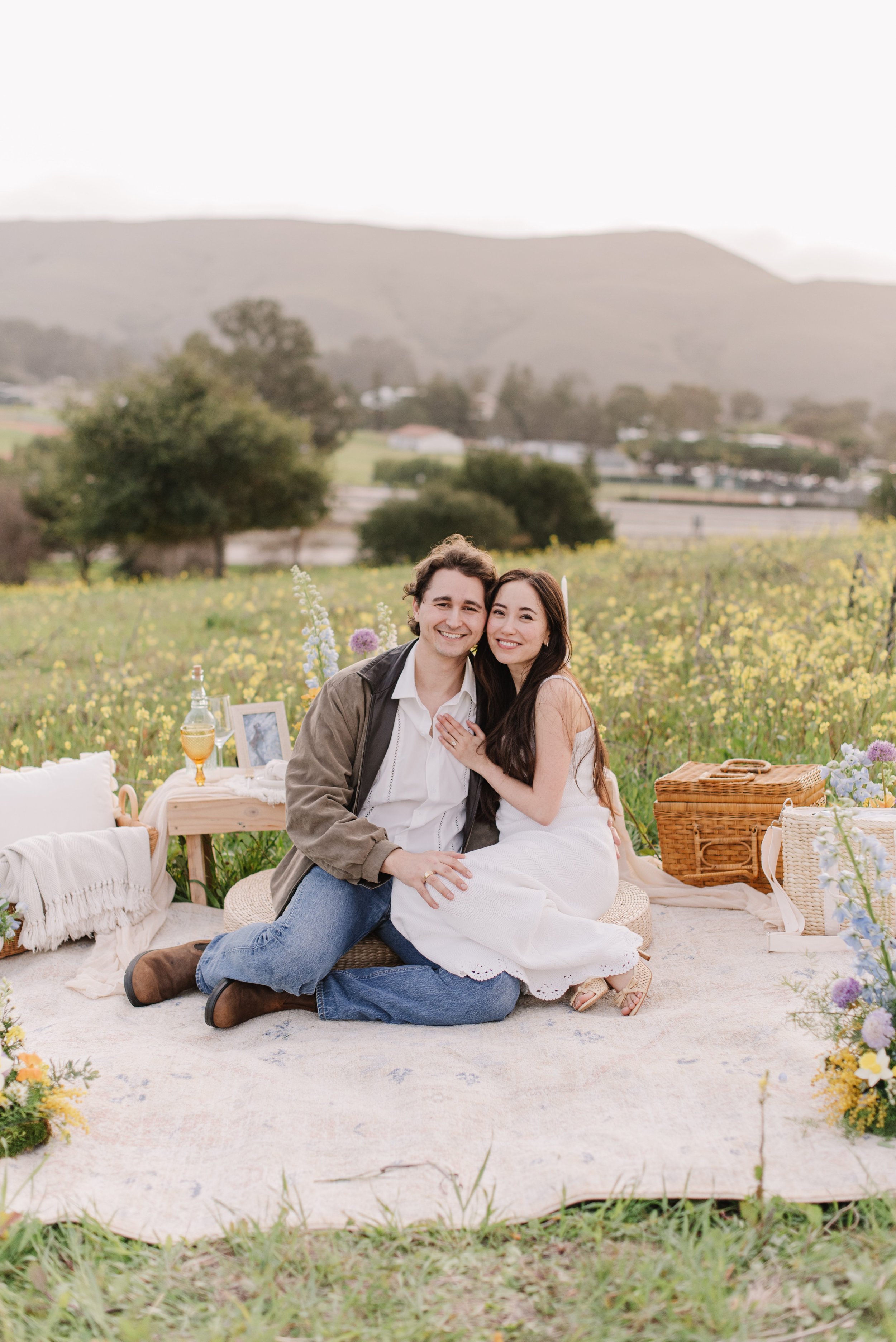 A happy couple sitting closely together on a picnic blanket outdoors with a scenic mountain background, surrounded by yellow wildflowers and greenery.