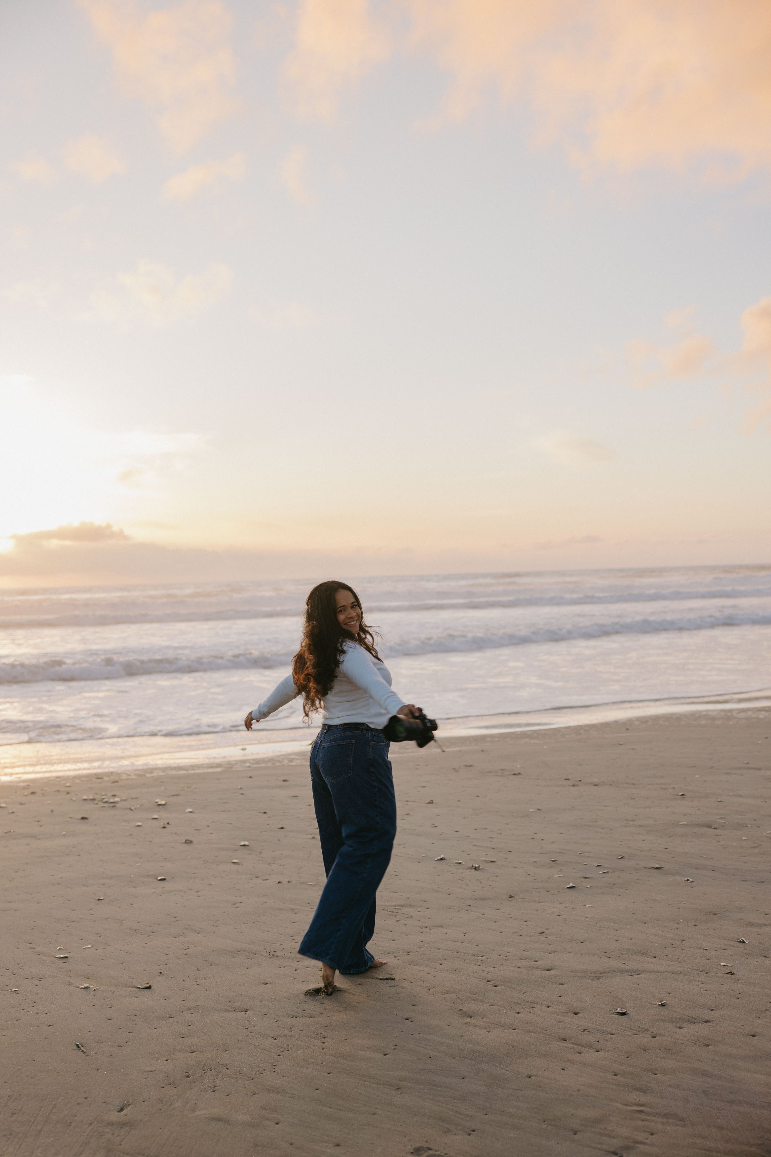 A woman with long dark hair smiling and holding a camera stands barefoot on a sandy beach with the ocean behind her at sunset.