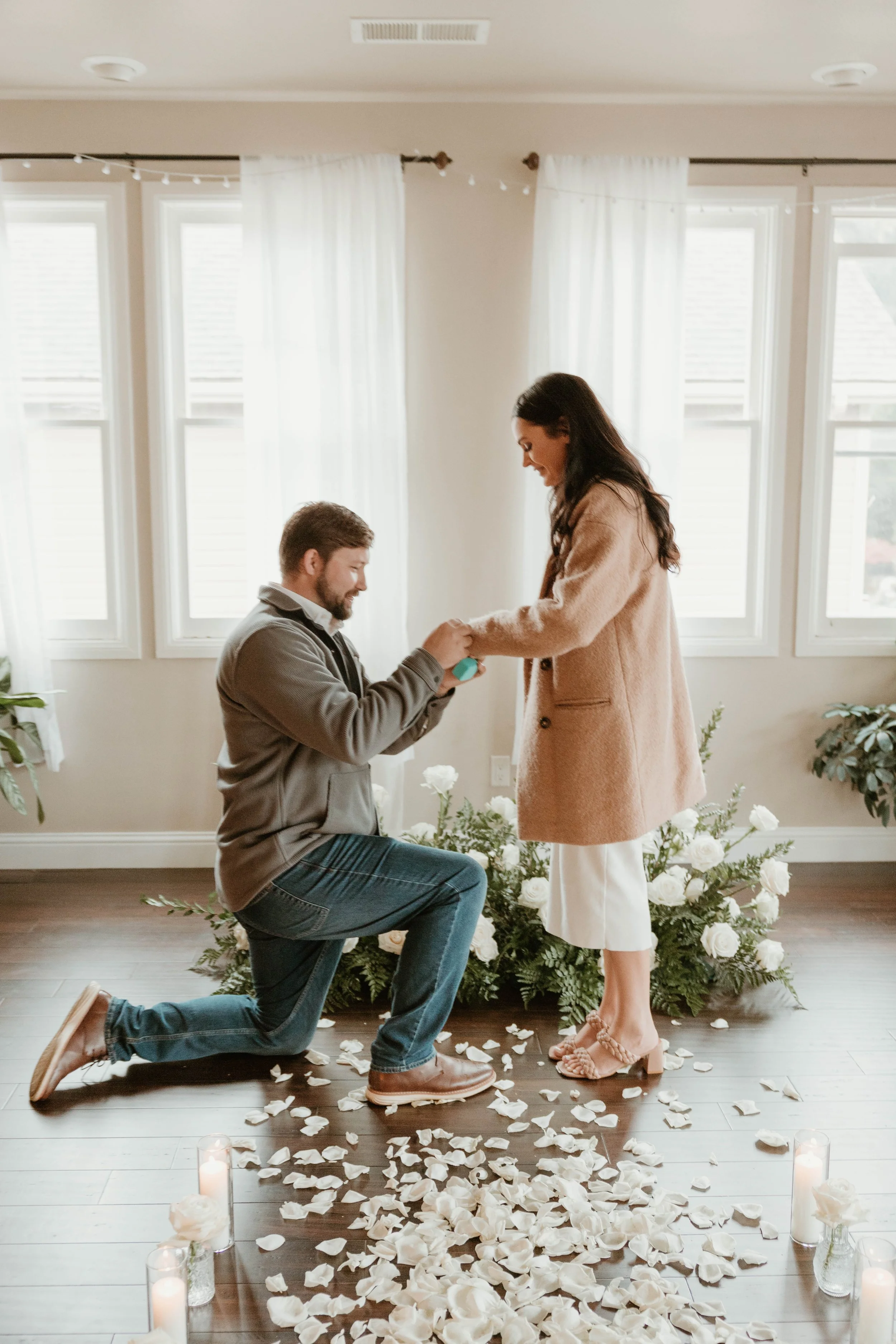 A man kneeling on one knee on a wooden floor, proposing with a ring to a woman standing in front of him, surrounded by white rose petals and candles, with large windows and white curtains in the background.
