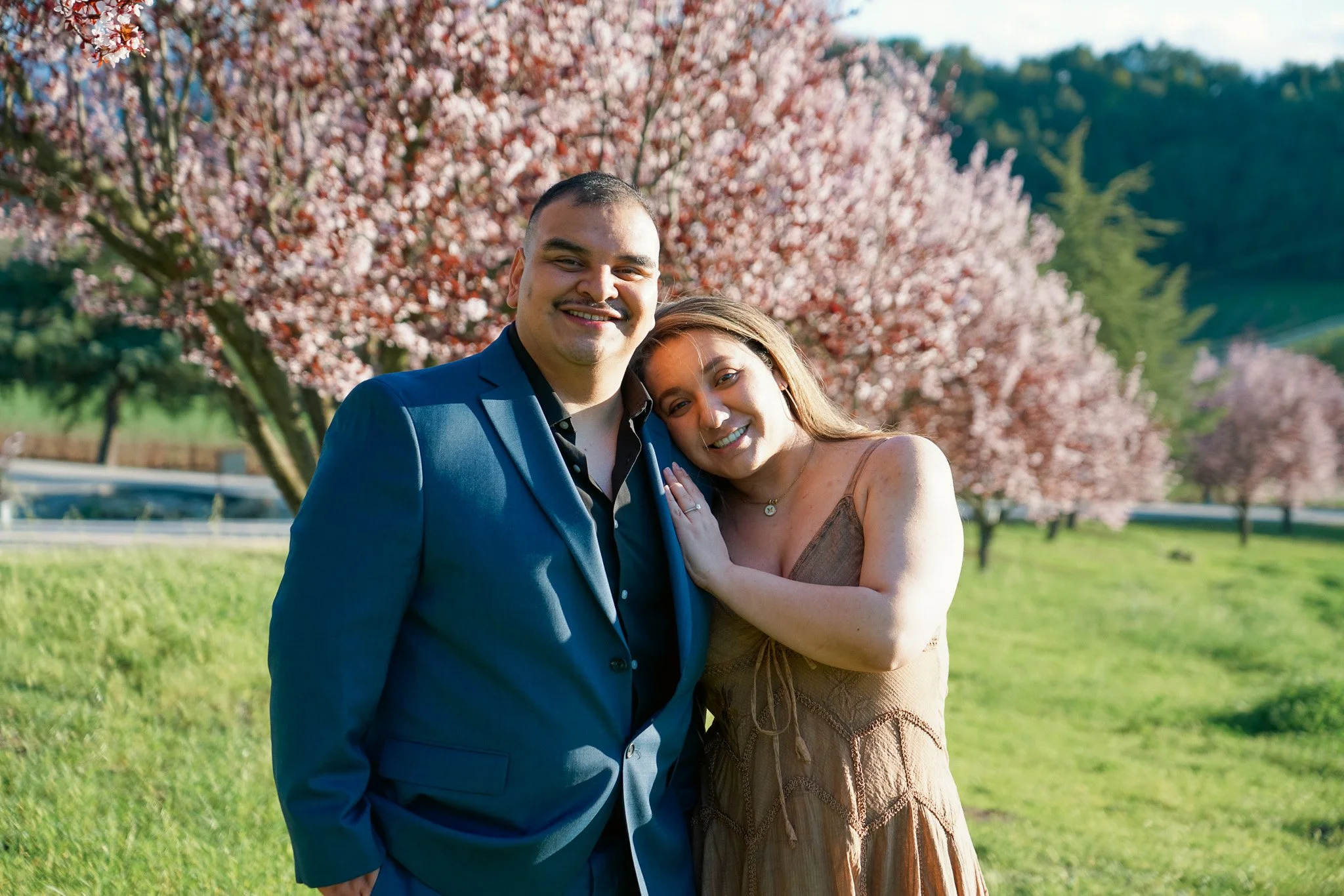 A couple posing outdoors in front of blooming pink spring trees, with the man wearing a blue suit and the woman in a brown dress, both smiling.