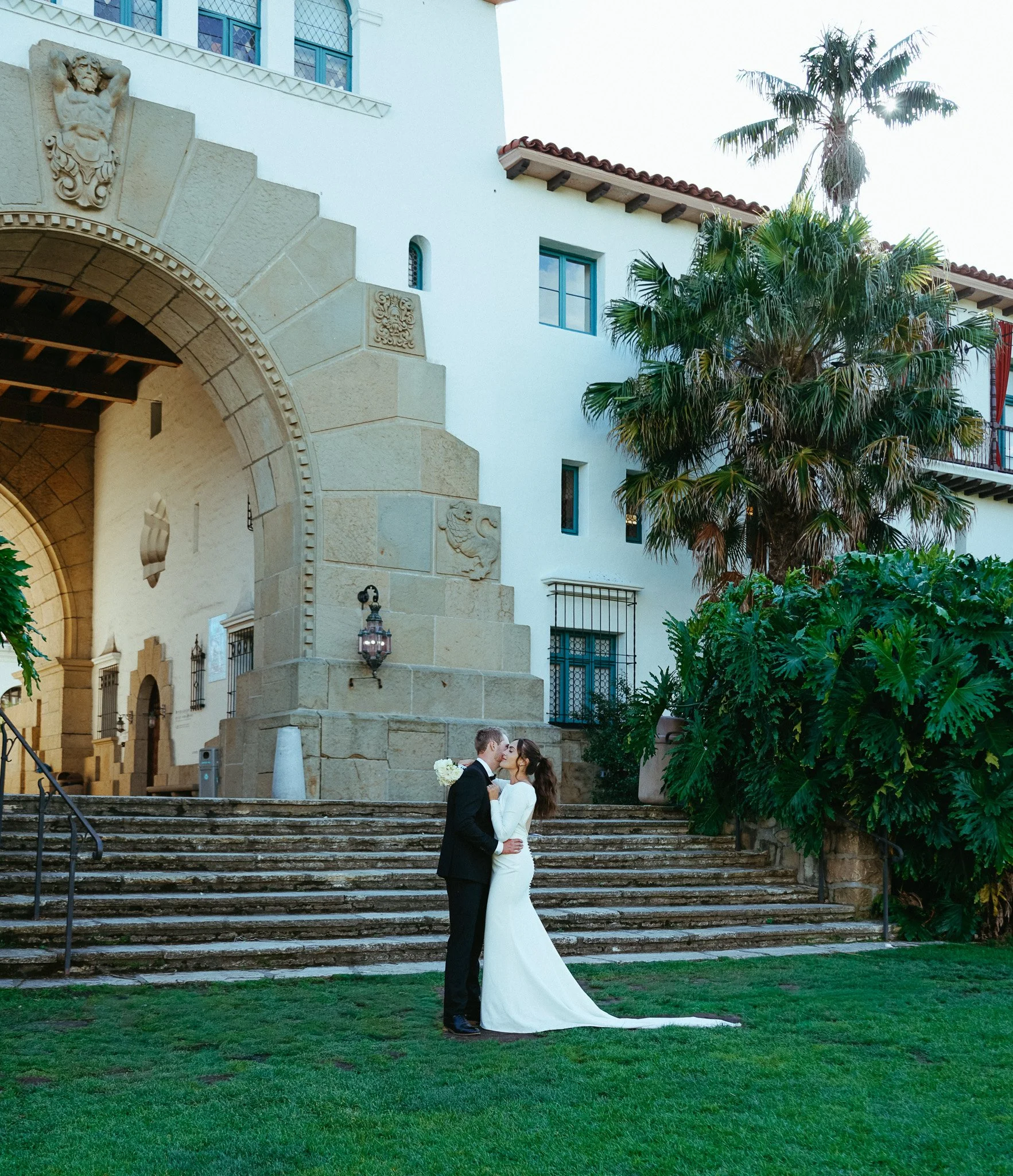 A bride and groom standing on a set of stone steps in front of a historic building with a large archway and decorative stone details. The bride wears a long white wedding dress and the groom wears a black suit. They are about to kiss, with the groom holding a bouquet of white flowers. Tall green palm trees are visible to the right, and the sky is bright with sunlight.