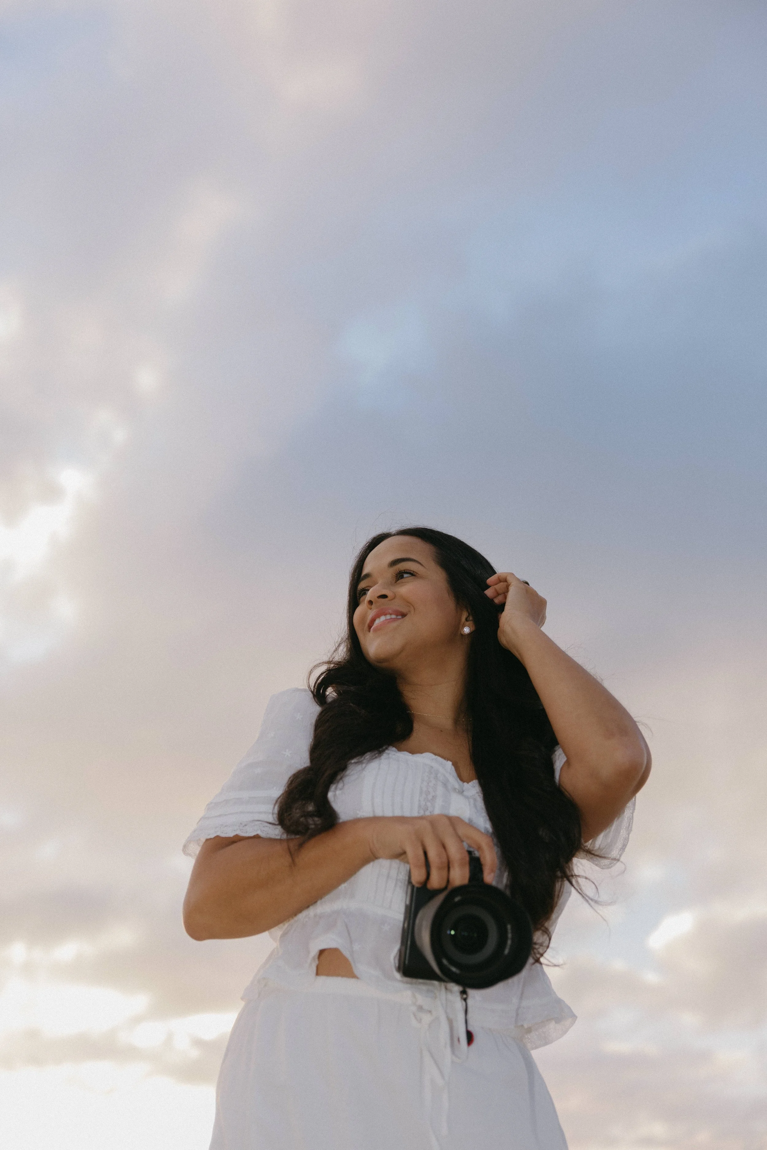 A woman with long dark hair wearing a white dress, smiling and holding a camera, standing outdoors against a cloudy sky.