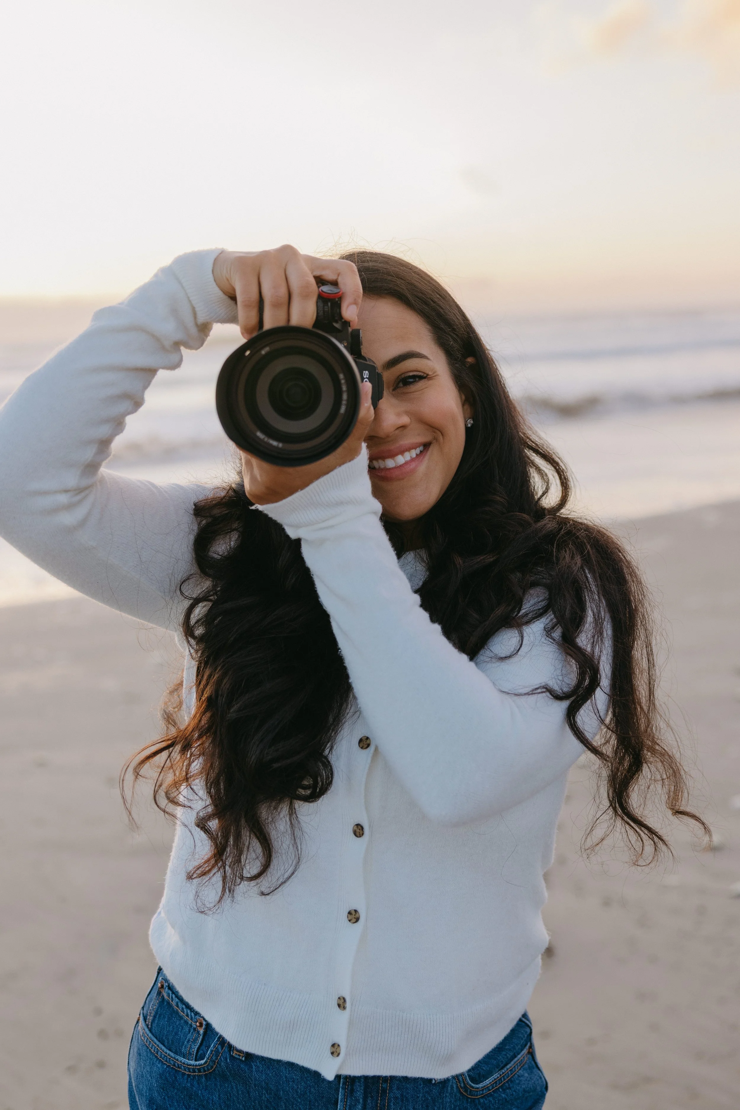A woman with long dark hair smiling on a beach during sunset, holding a camera and taking a photo.