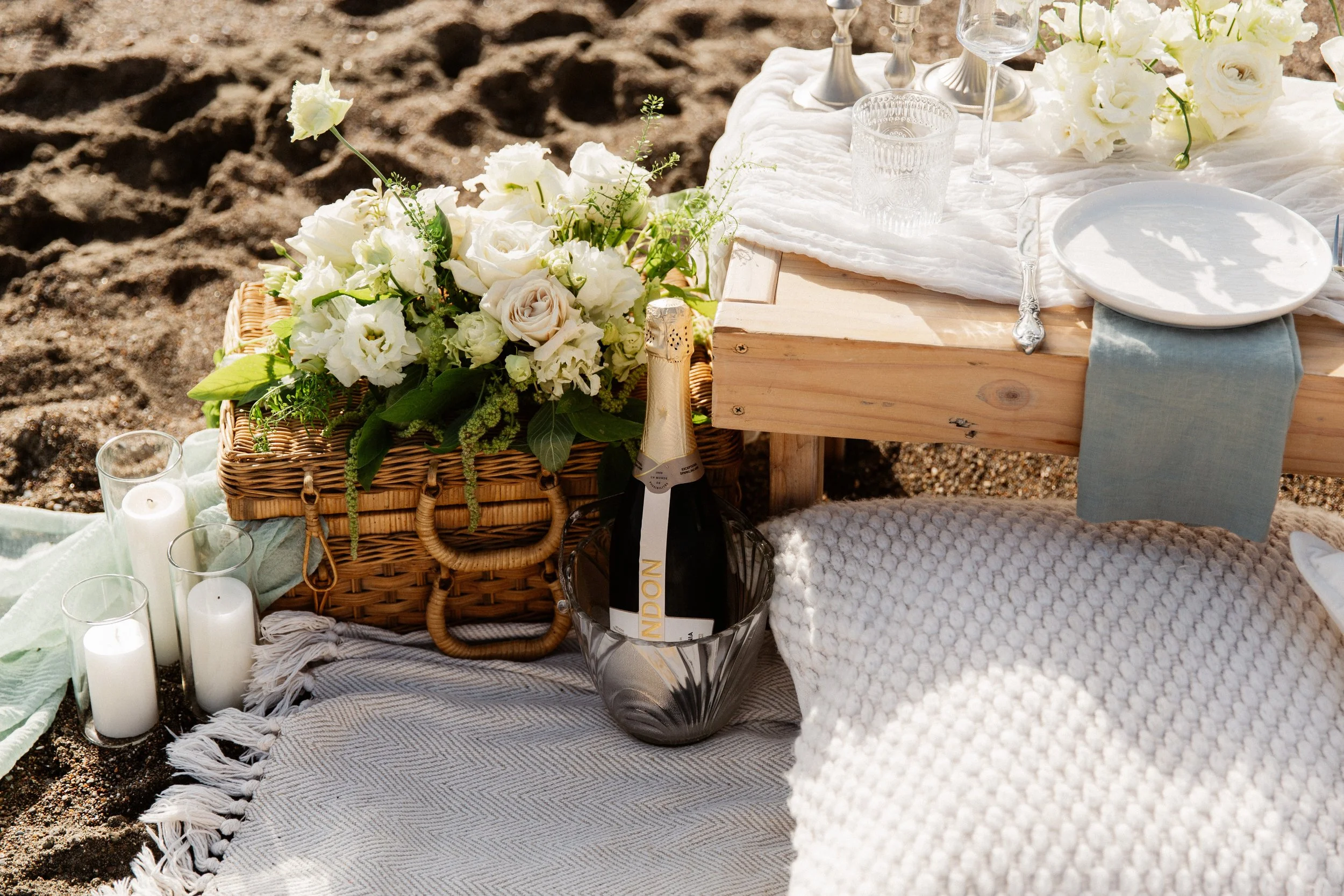 A picnic setup on sandy ground featuring a basket of white and cream flowers, candles, empty glasses, and a bottle of champagne, with a wooden table and white tableware.