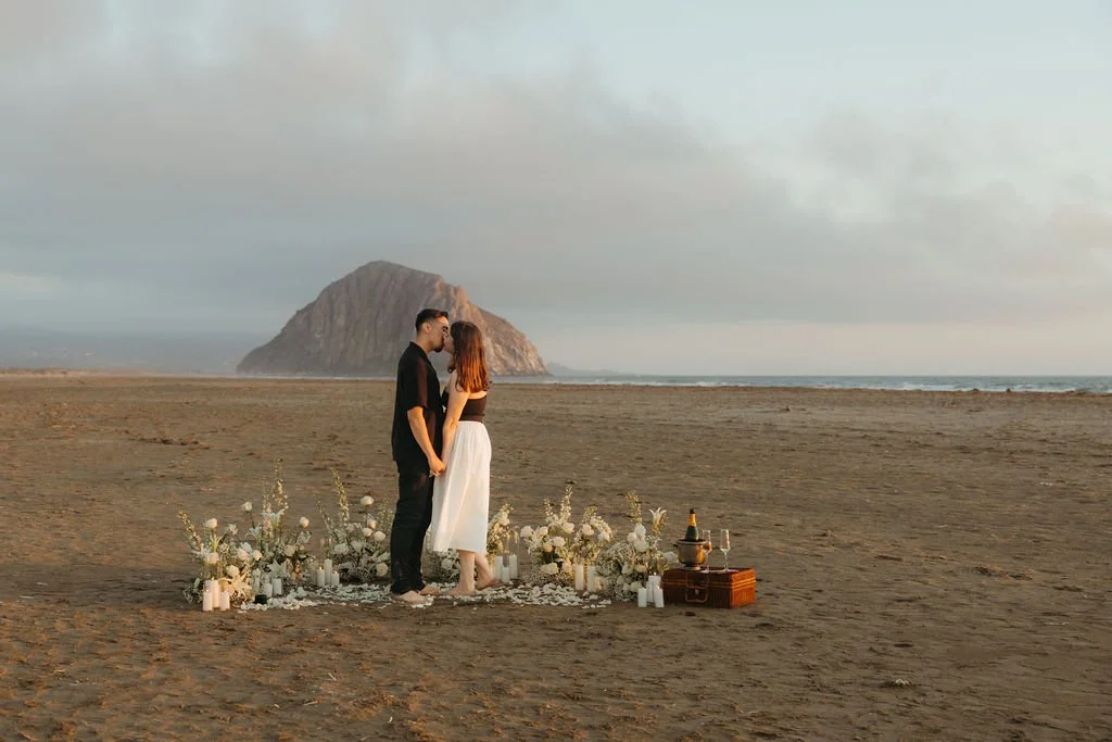 A couple standing close on a decorated beach during sunset, with large floral arrangements, candles, and a vintage camera on a box nearby, and a large rock formation in the background.